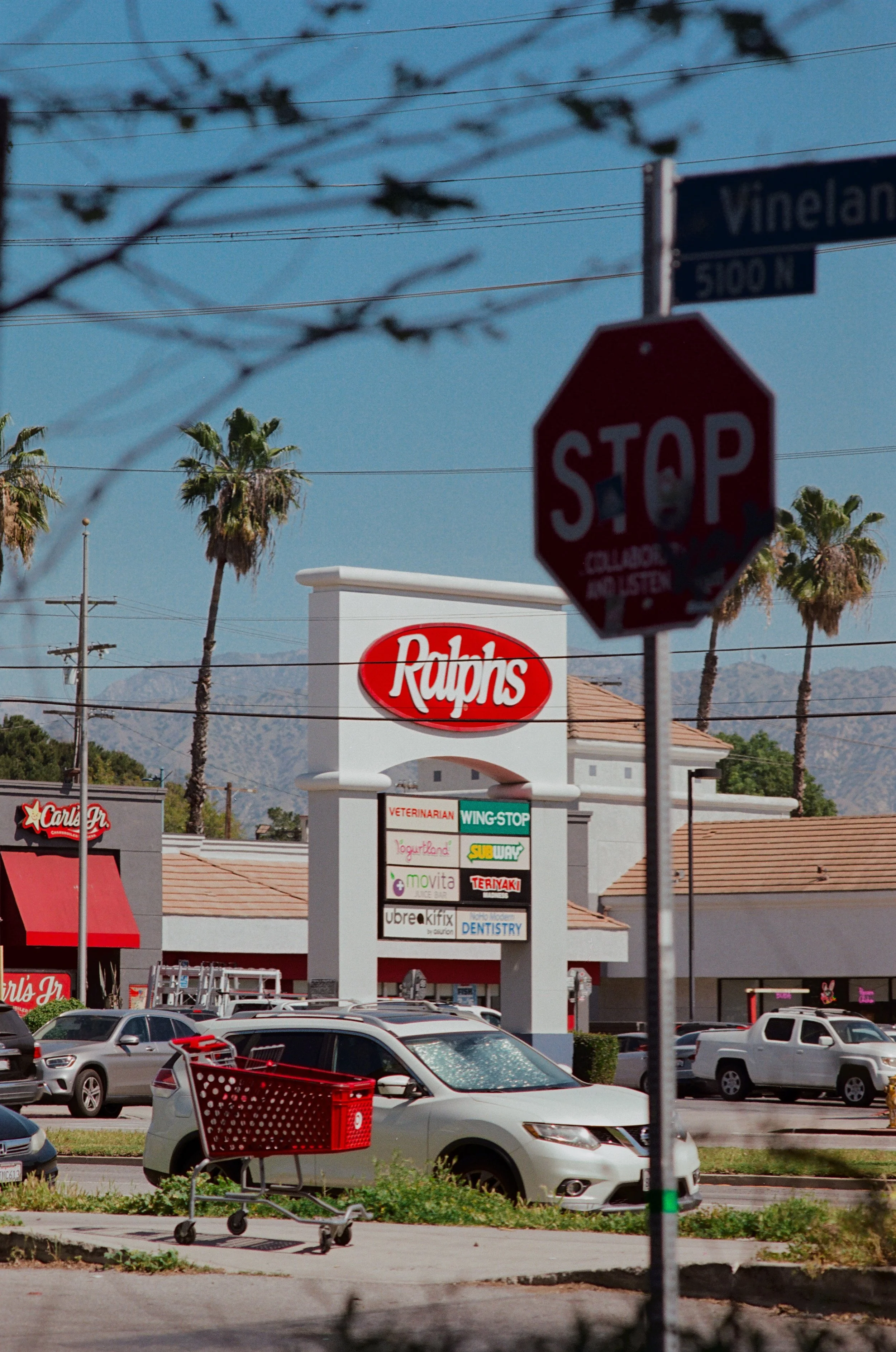 A parking lot filled with cars in front of a shopping center, with palm trees, a stop sign, traffic lights, power lines, and a billboard showing various store signs, with mountains visible in the background.