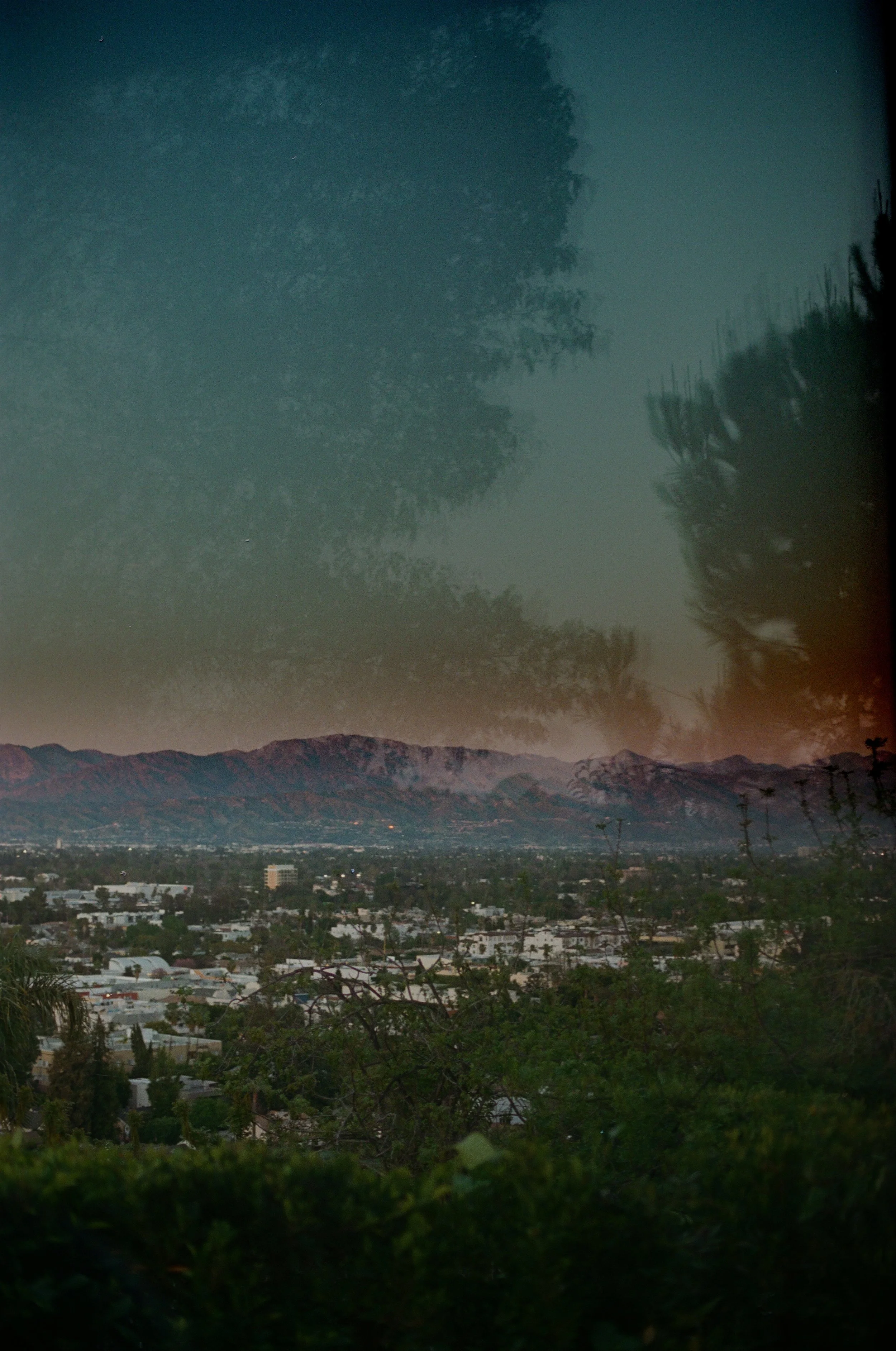 A landscape view of a town with mountains in the background at dusk, and a reflection of trees and the sky in a body of water in the foreground.