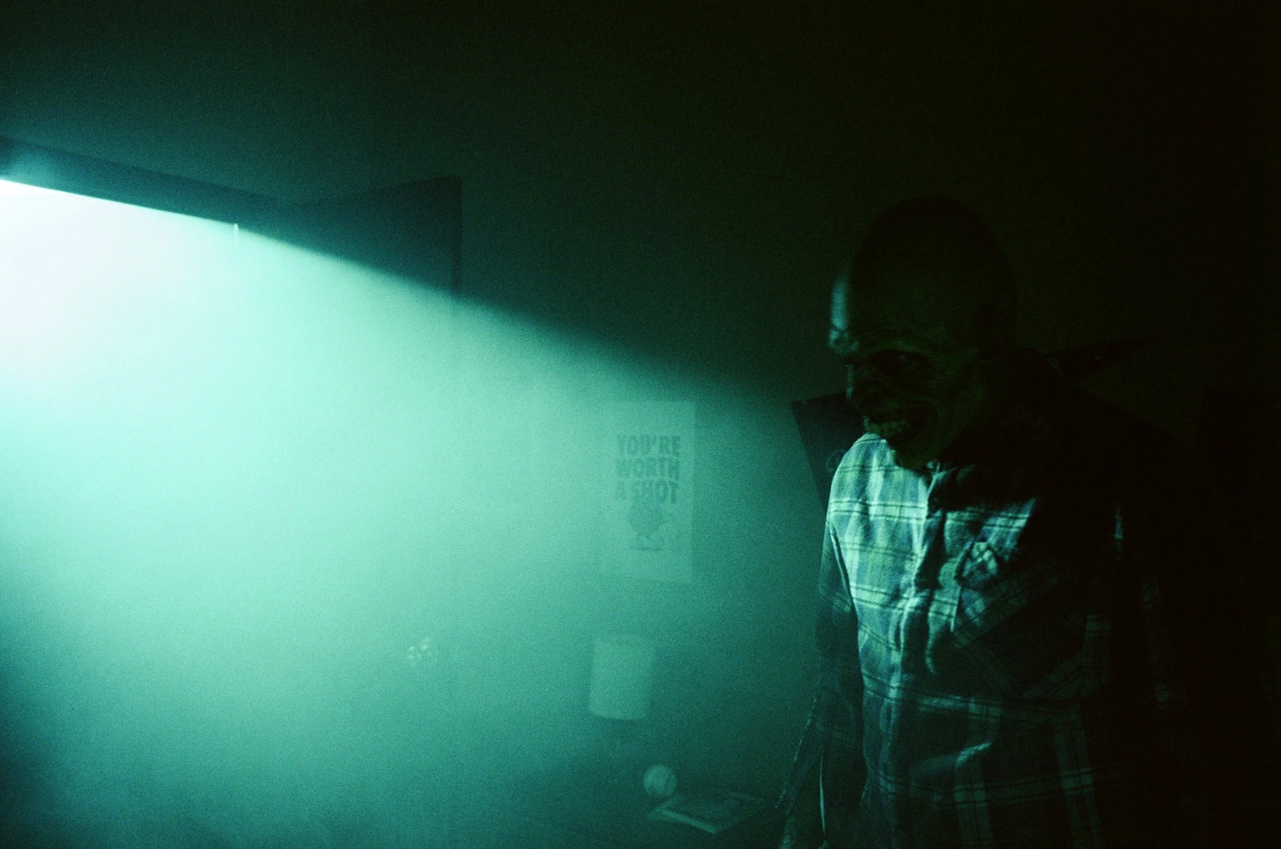A person in a hooded sweatshirt and checkered shirt standing in dimly lit room with green and blue lighting.