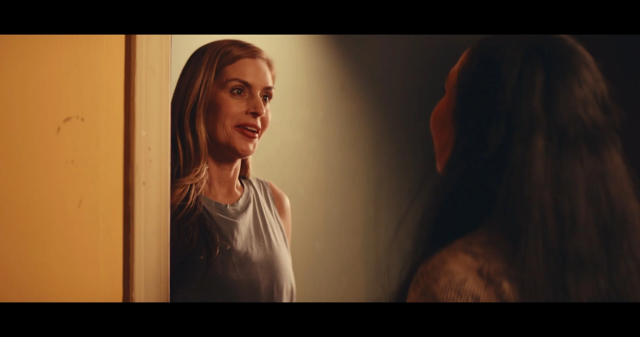 A woman with long, wavy brown hair and a sleeveless gray top looks at another person with long hair, standing in a doorway with yellow walls.