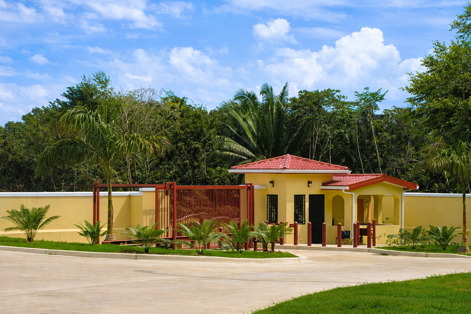 A small yellow house with a red-tiled roof, surrounded by tropical plants and trees, with a concrete driveway and a clear blue sky overhead.