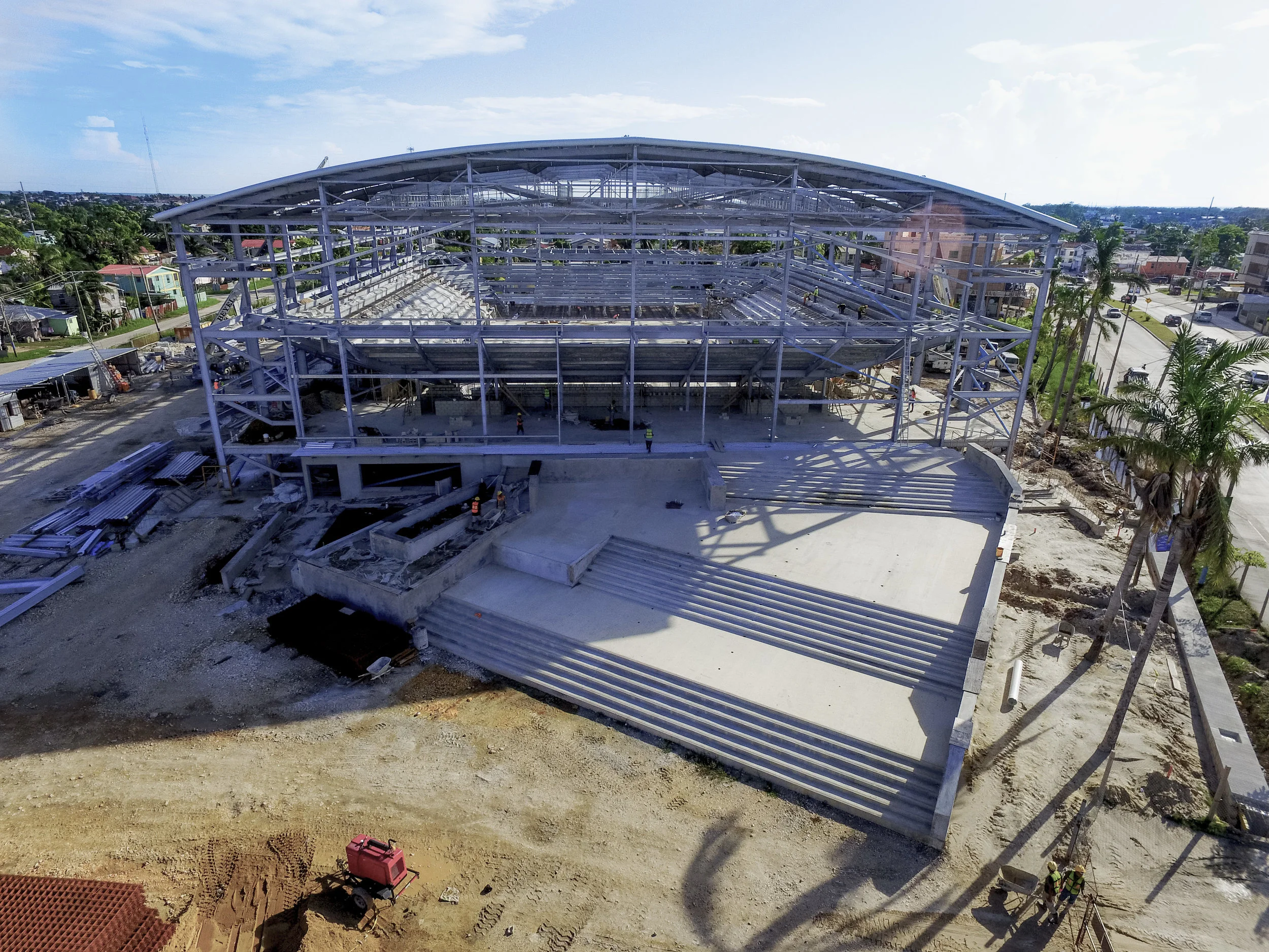 Construction site with a large metal framework of a building under construction. Several workers are working on the structure, and construction materials are visible around the site. Residential houses and trees are in the background under a partly c