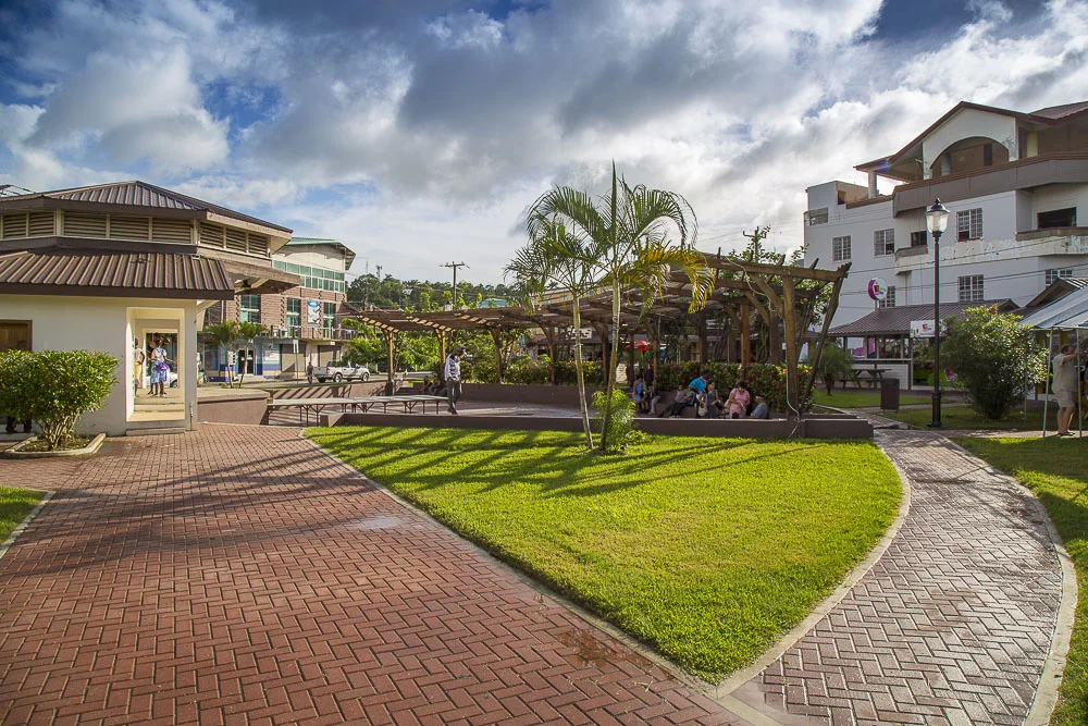 A park with a brick walkway, a grassy area, and a shaded sitting area with people. There are buildings around the park, palm trees, and a partly cloudy sky.
