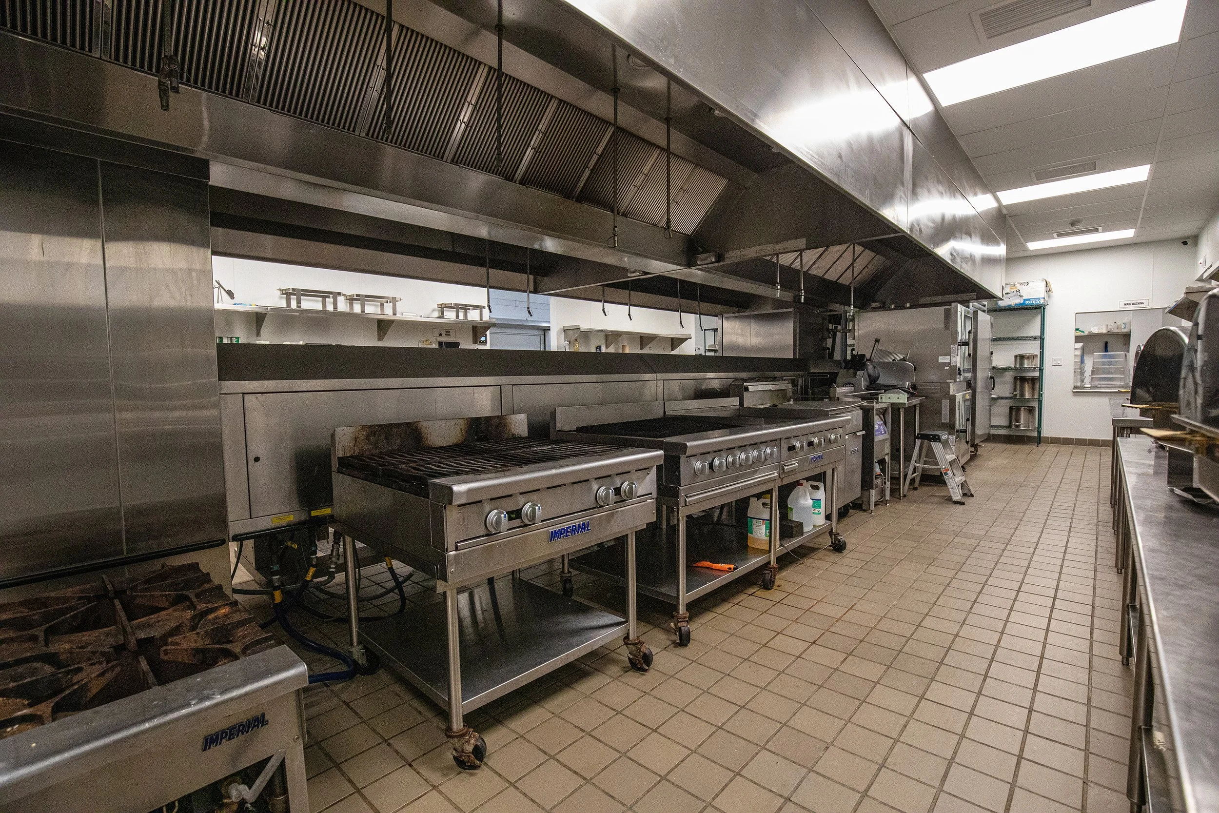 Commercial kitchen with stainless steel appliances, including stoves, ovens, and a large ventilation hood, tiled floor, and shelves in the background.