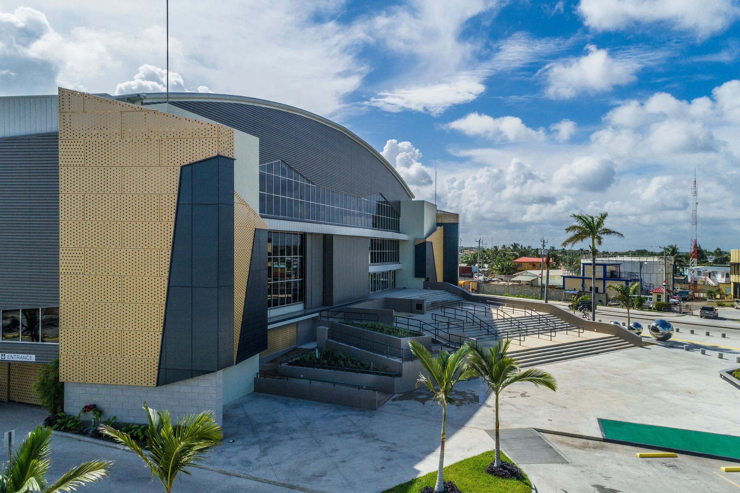 Modern building with curved roof, glass windows, and gold perforated panels, surrounded by palm trees and a parking lot, under a partly cloudy sky.