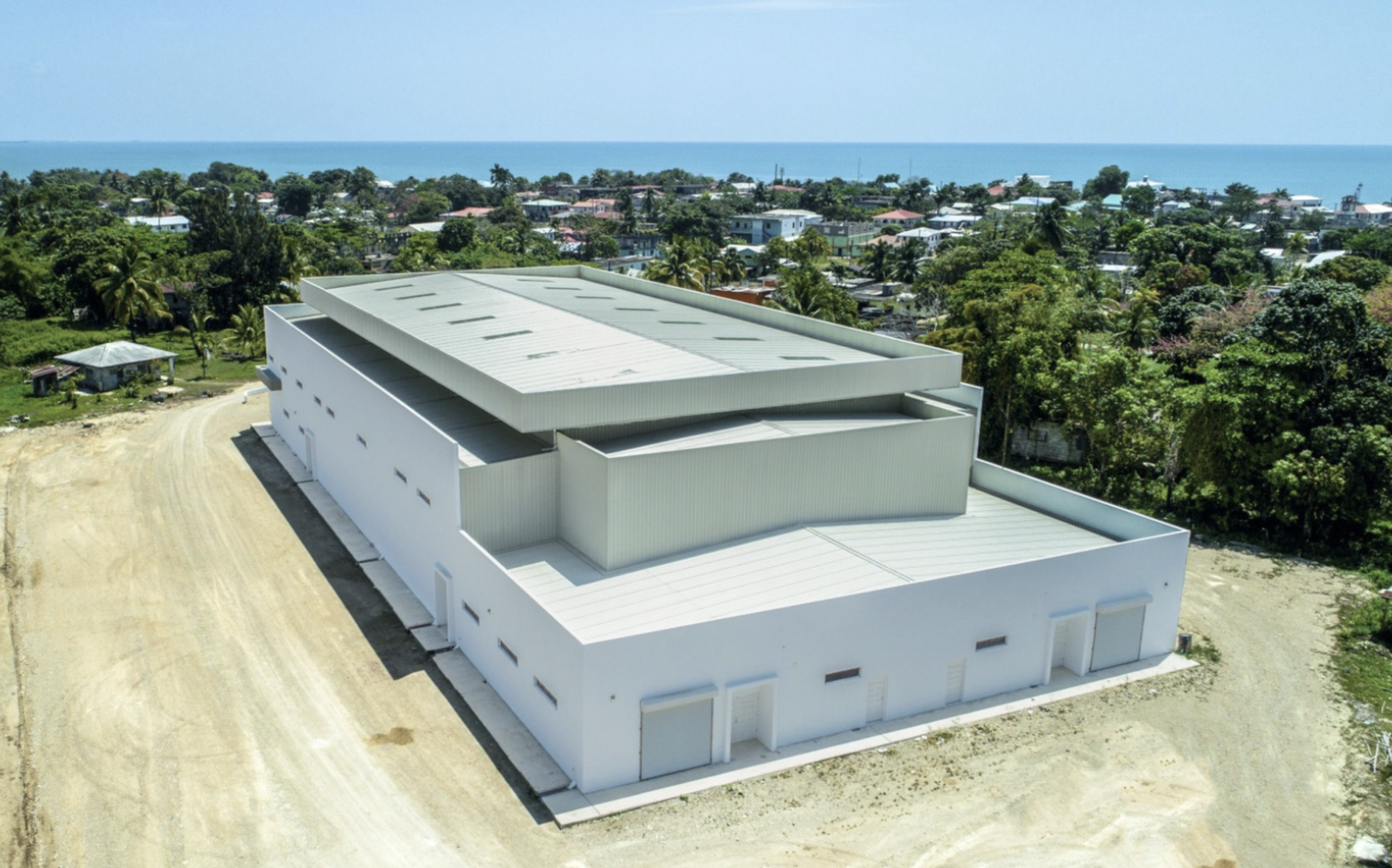 Aerial view of a modern white building with a metal roof, situated near a coastal town with trees and houses, and the ocean in the background.