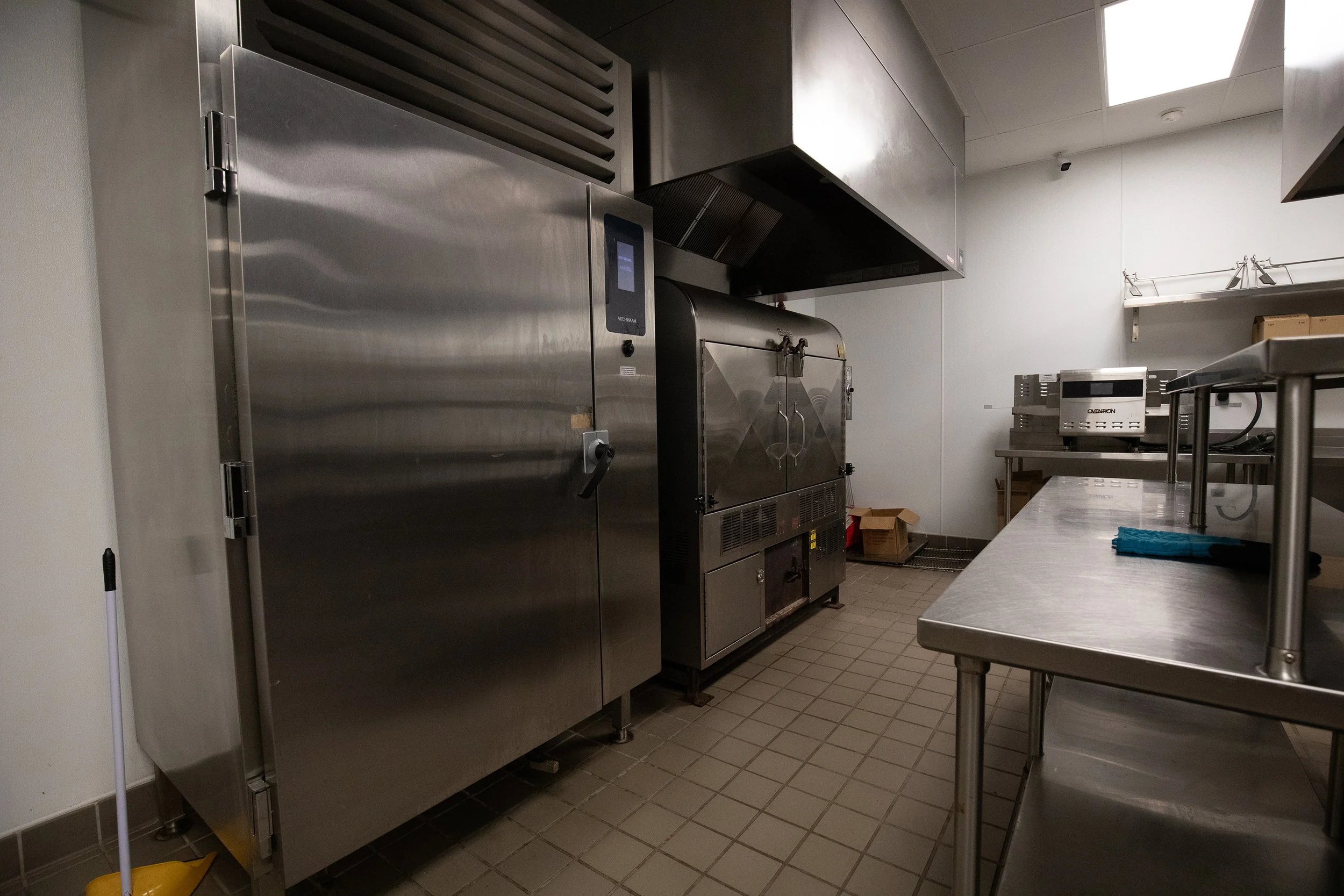 Commercial kitchen with stainless steel appliances, including an oven and a large refrigerator, a work table, and some cardboard boxes on the floor.