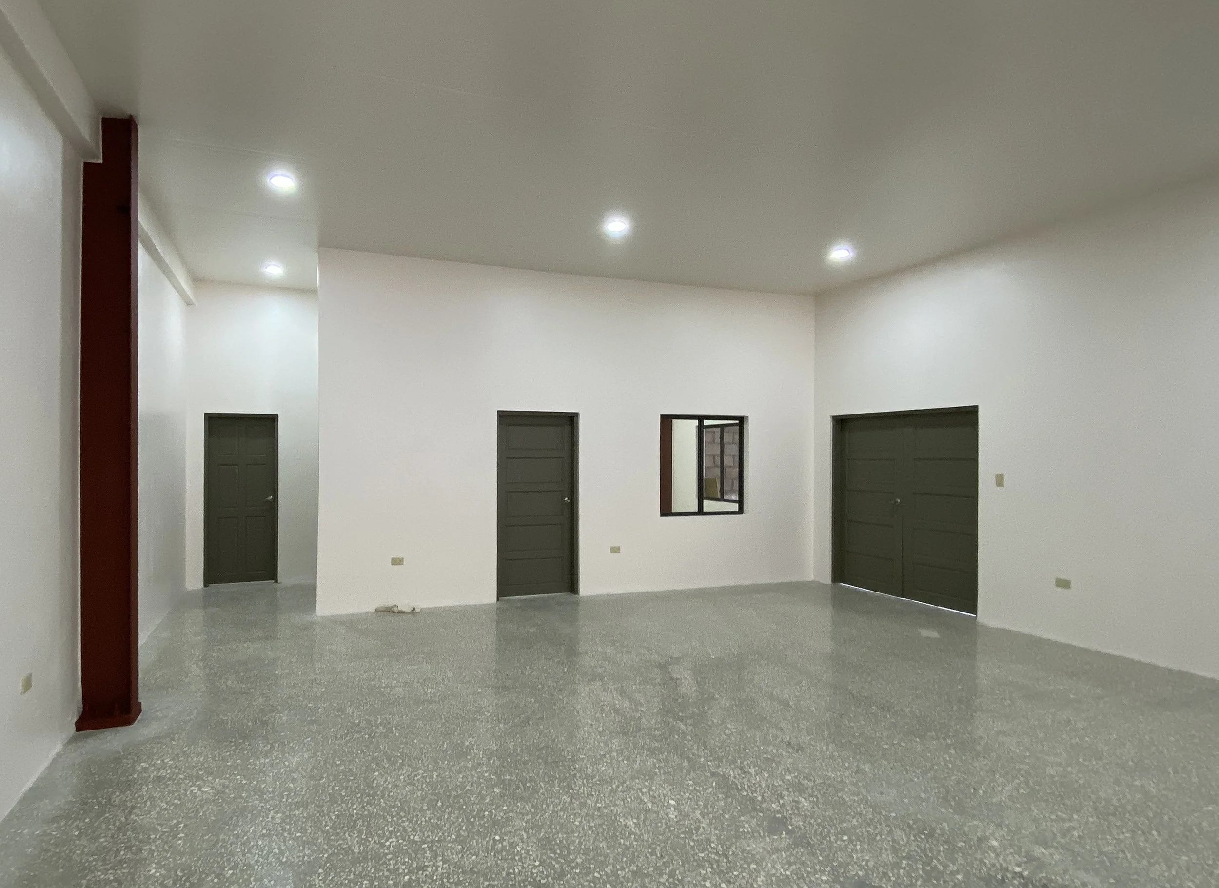Empty room with white walls, gray speckled floor, and several closed gray doors, with small windows in some doors and a mirror on one wall.