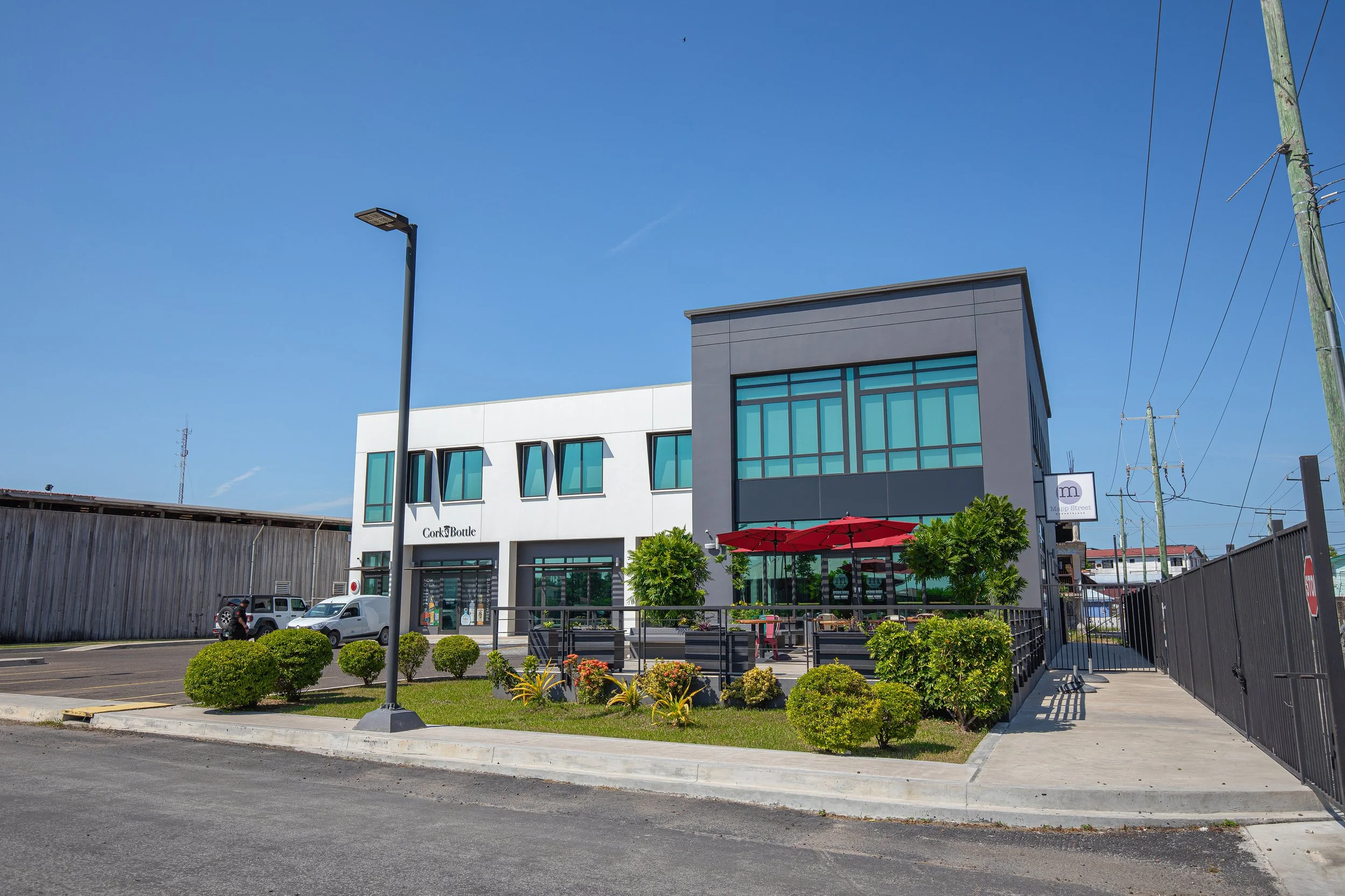 Modern building with large windows, outdoor seating with red umbrellas, green landscaping, parked cars, street lamp, and utility poles under a clear blue sky.