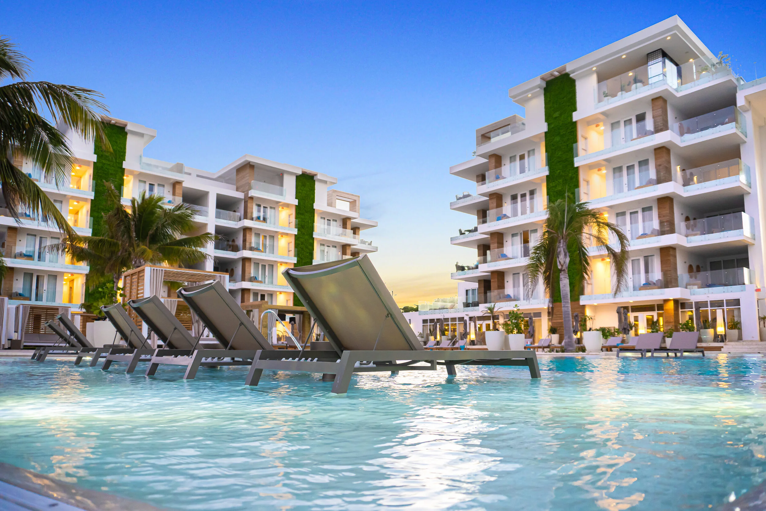 Luxury hotel pool area with lounge chairs and a modern resort building in the background during sunset.