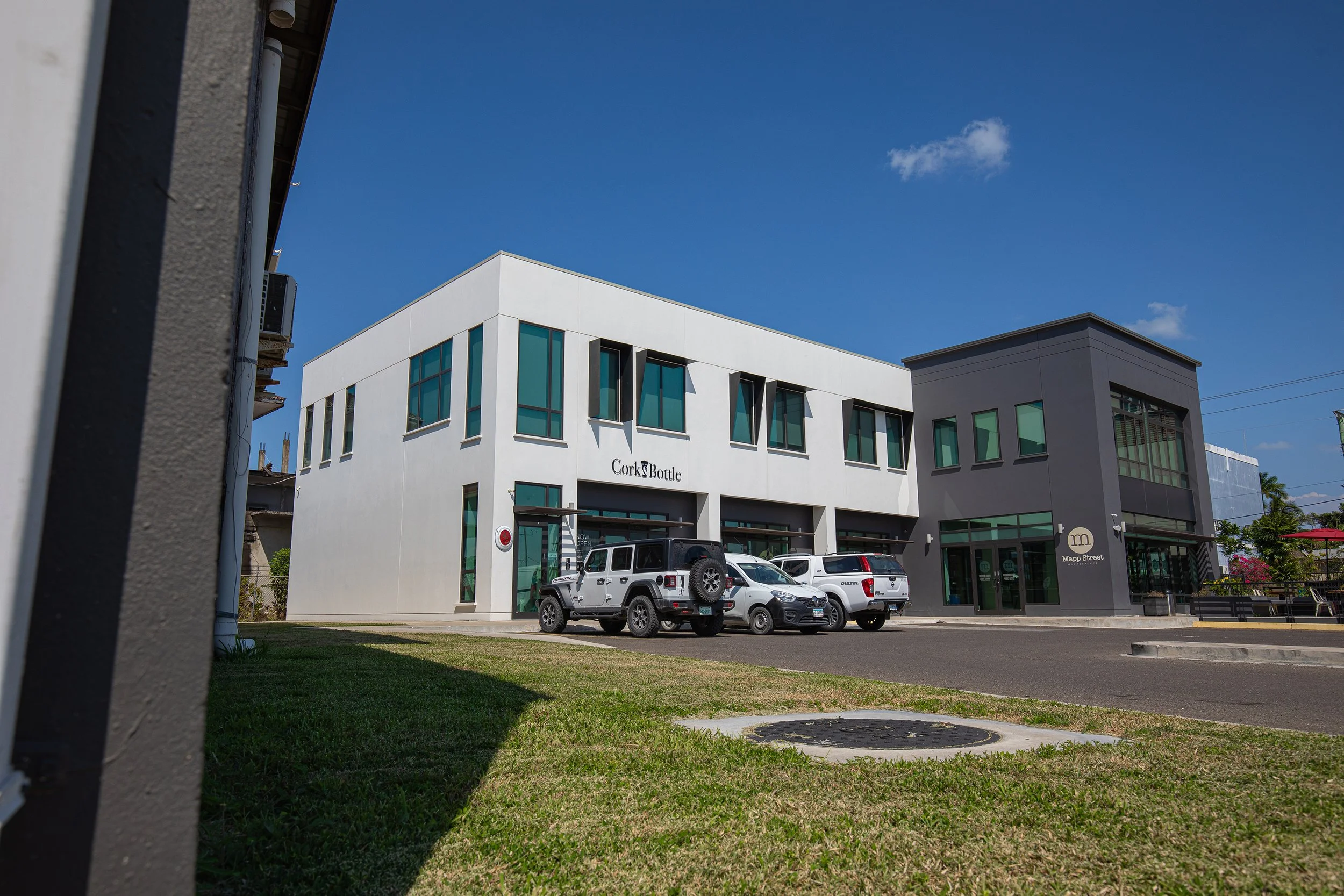 Street view featuring modern white and gray commercial buildings with parked cars in front, including a Jeep, with a blue sky and small clouds overhead.