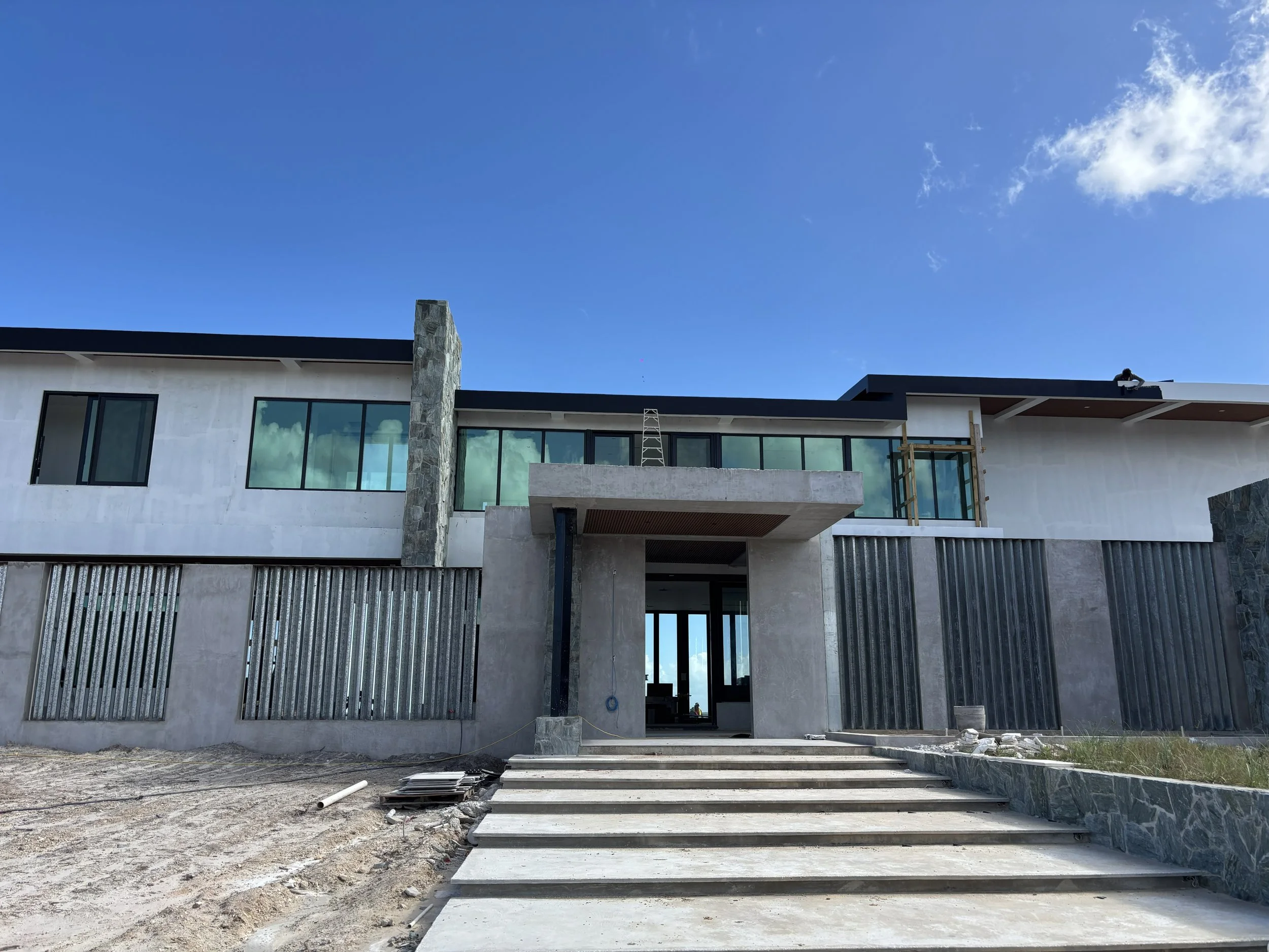 Modern house under construction with a staircase leading to the entrance, large windows, and a clear blue sky in the background.