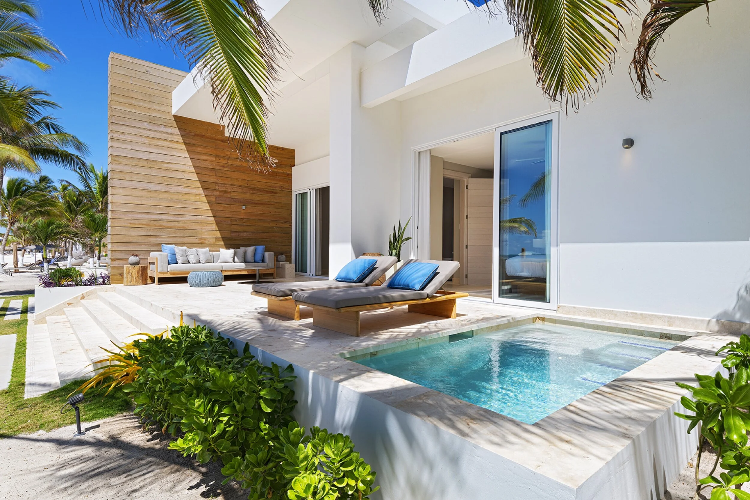 Modern beach house with outdoor patio, lounge chairs, swimming pool, and tropical palm trees under a clear blue sky.