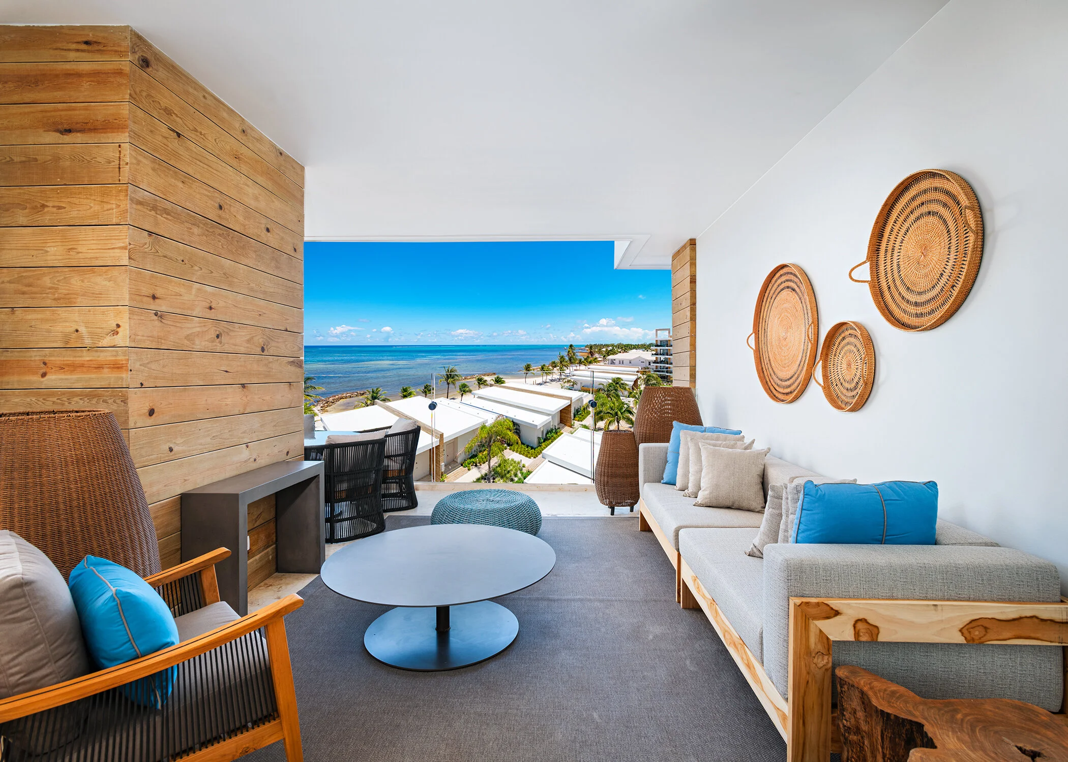 A balcony view with a white sofa decorated with blue and beige pillows, a round coffee table, brown woven chairs with pillows, a wall with three woven baskets, a wooden accent wall, and an ocean view with white buildings and palm trees under a bright