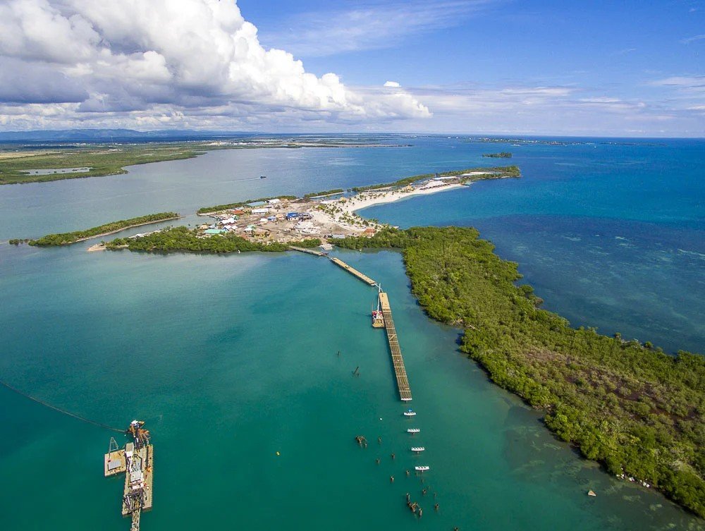 An aerial view of a coastal area with turquoise waters, a breakwater extending into the ocean, a small town or village with buildings, green vegetation, and sandy beaches under a partly cloudy sky.
