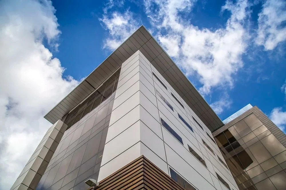 Low-angle view of a modern, multi-story building with white panels and glass windows, set against a blue sky with scattered clouds.