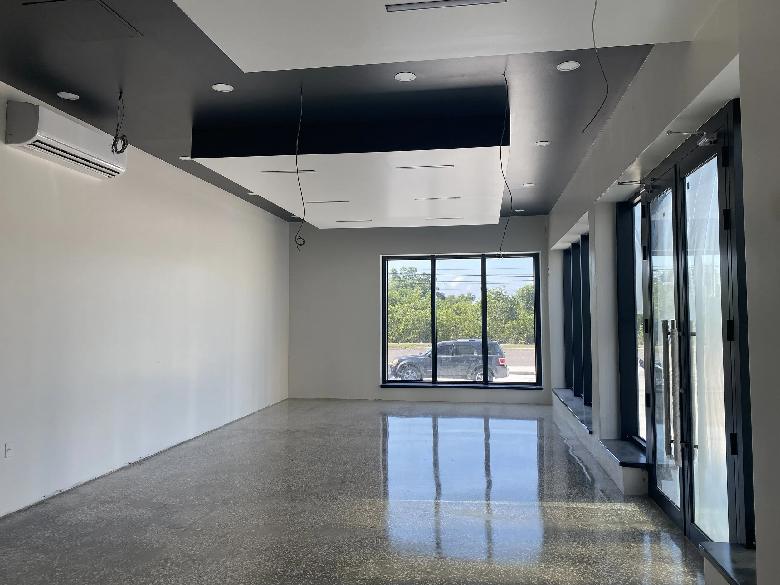 Empty commercial or office space with large windows, polished concrete floors, and a black ceiling with hanging wires and recessed lighting.