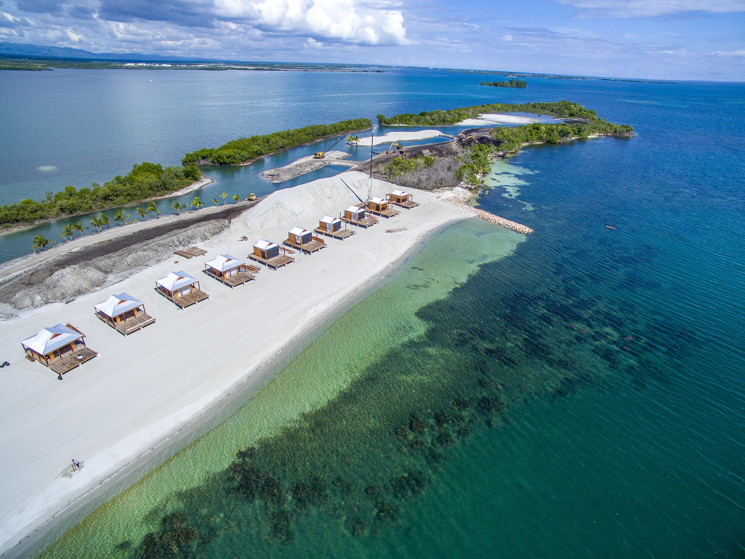 Aerial view of a construction site on a beach, with small huts, construction equipment, and a canal, surrounded by water and greenery.