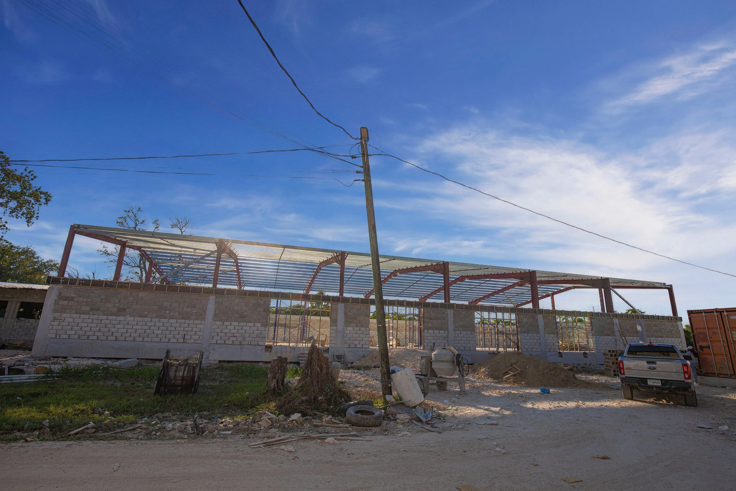 Construction site with a partially built brick structure, construction materials, a utility pole with multiple power lines, a car parked on dirt, trees, and a clear blue sky with some clouds.
