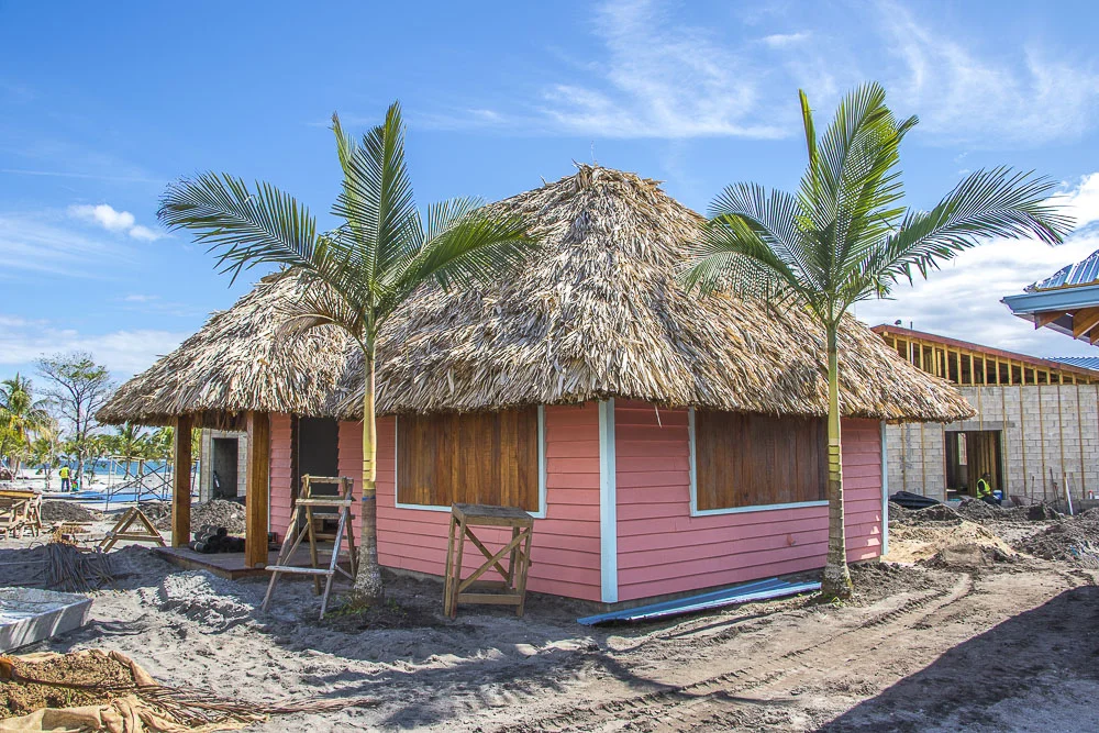 A pink house with a thatched roof under construction, surrounded by sand, palm trees, and construction materials.