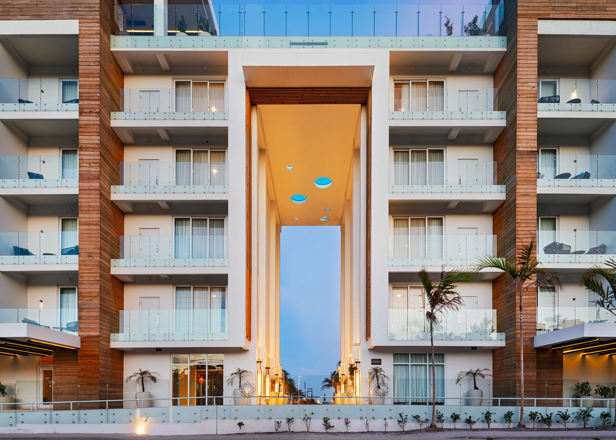 A modern multi-story residential building with balconies, glass railings, and wooden accents. The building has a central open passage with circular ceiling lights and decorative palm trees outside.