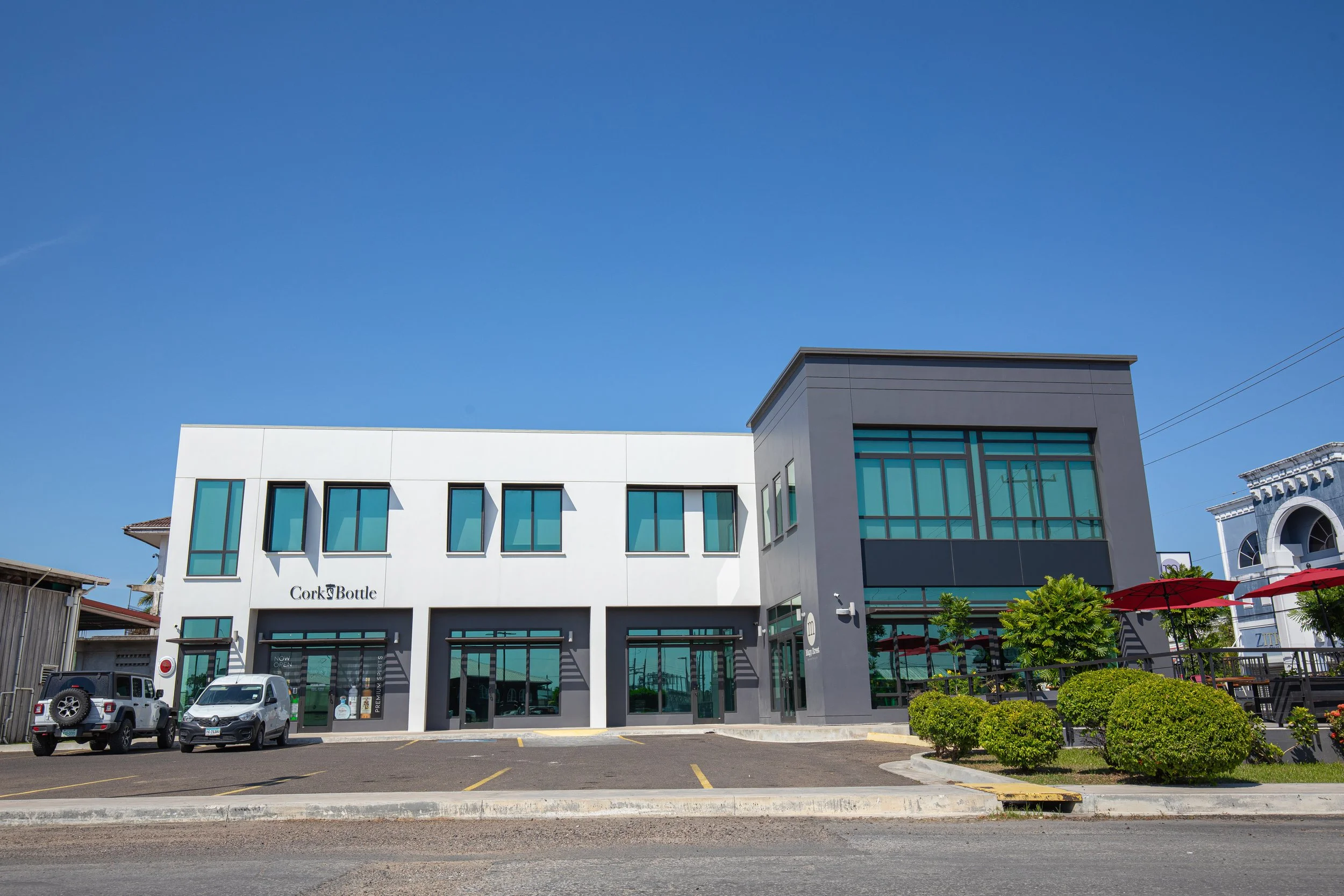 A modern white building with large windows and the name Cork Bottle on the front, with cars parked in front and a clear blue sky overhead.