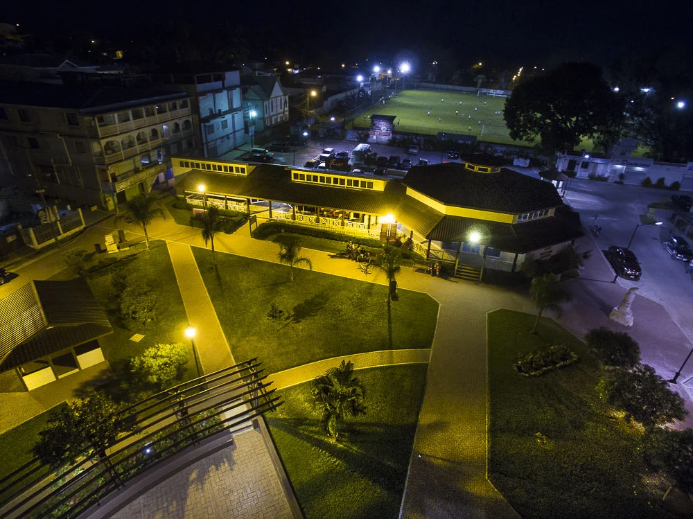 Nighttime aerial view of a small park with walkways, trees, and benches, illuminated by yellow streetlights, with a building and a sports field with lights in the background.