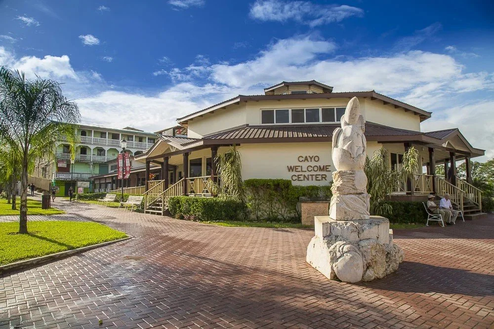 A building labeled 'Cayo Welcome Center' with stone sculpture in front, brick pathway, benches, palm trees, and a blue sky with clouds.