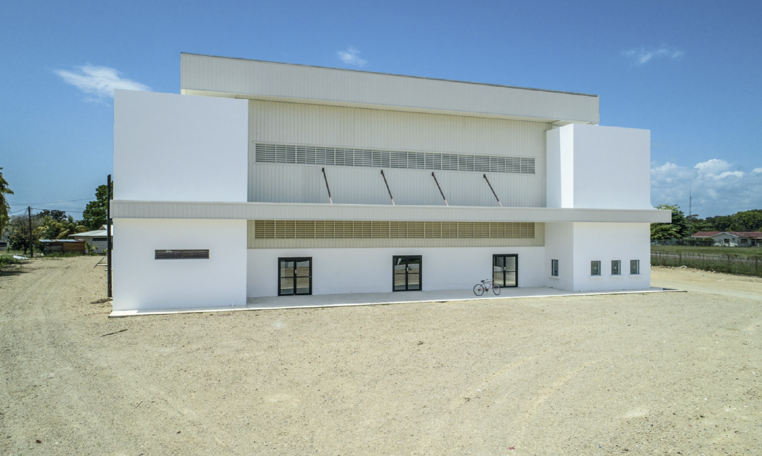 Modern white building with flat roof and multiple windows, bicycle parked outside, on sandy ground under a blue sky.