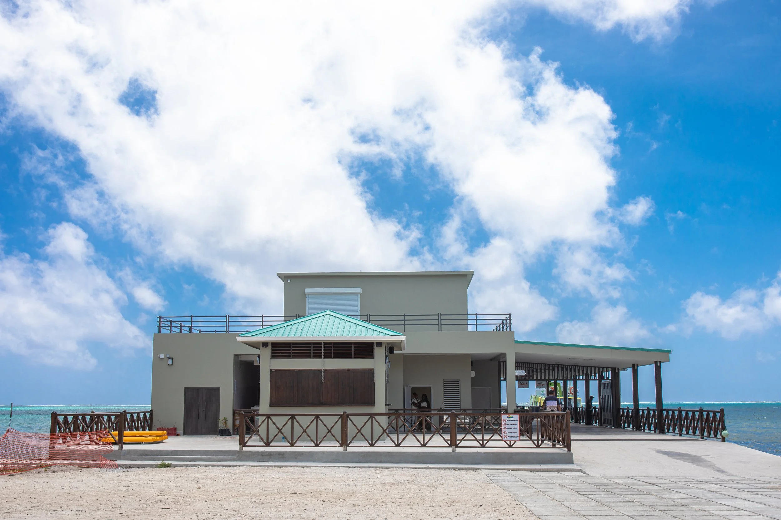 Beachside building with a turquoise roof, on a sandy area with a wooden fence, under a partly cloudy sky.