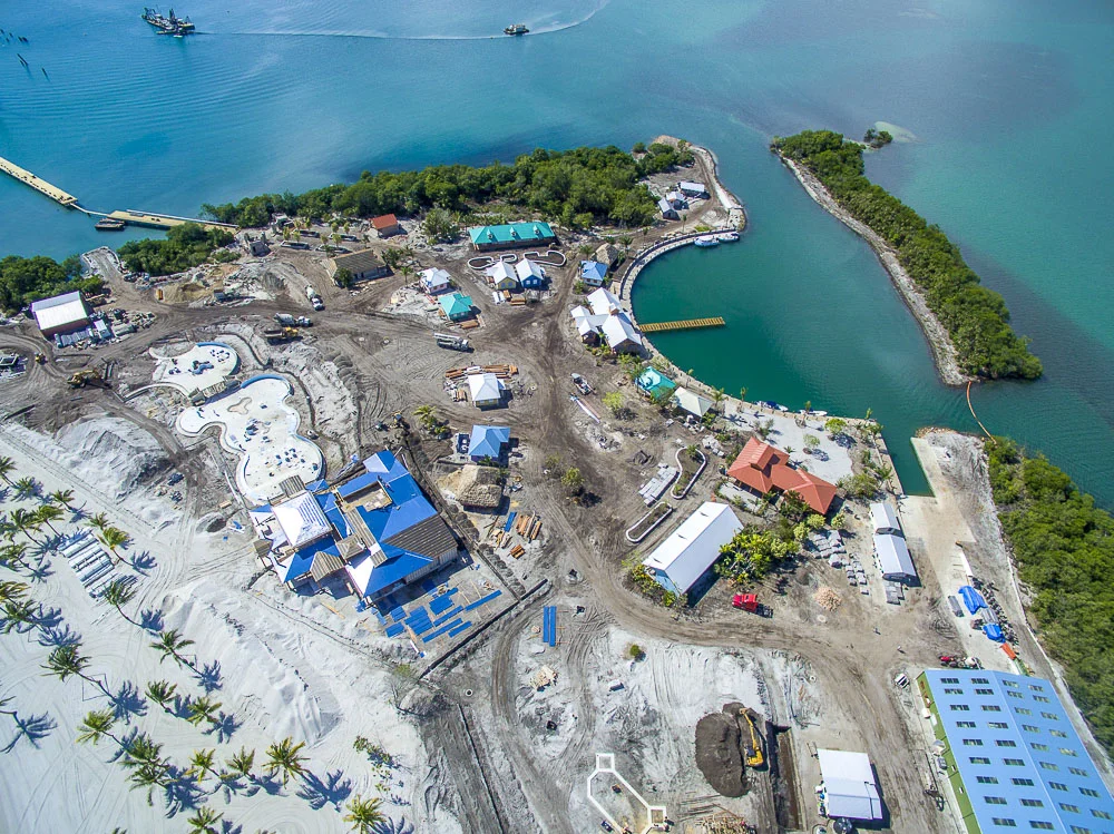 Aerial view of a construction site near water with buildings, swimming pools, and a marina.