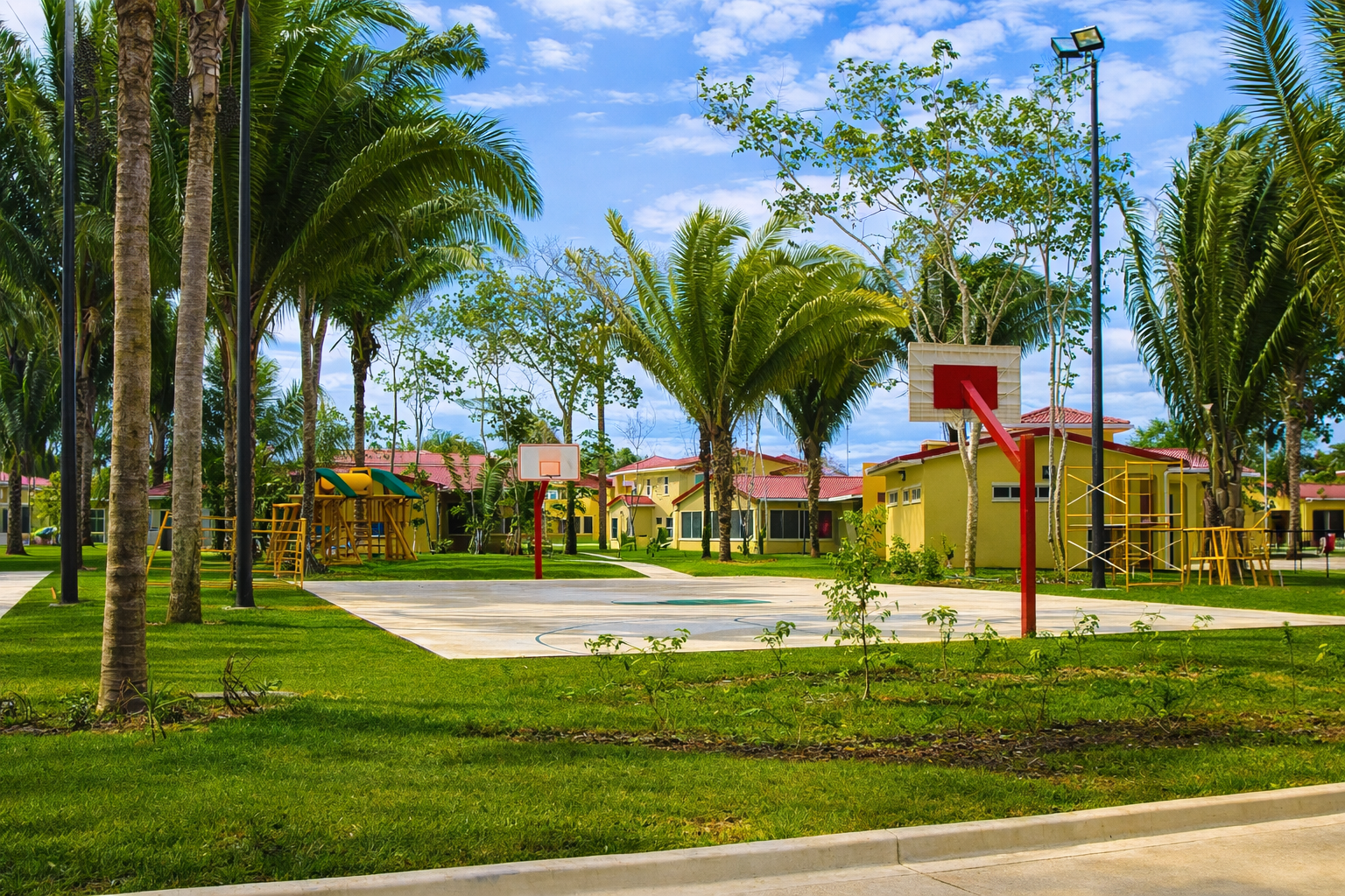 Empty outdoor basketball courts with two hoops, surrounded by palm trees, yellow houses, and playground equipment under a partly cloudy sky.
