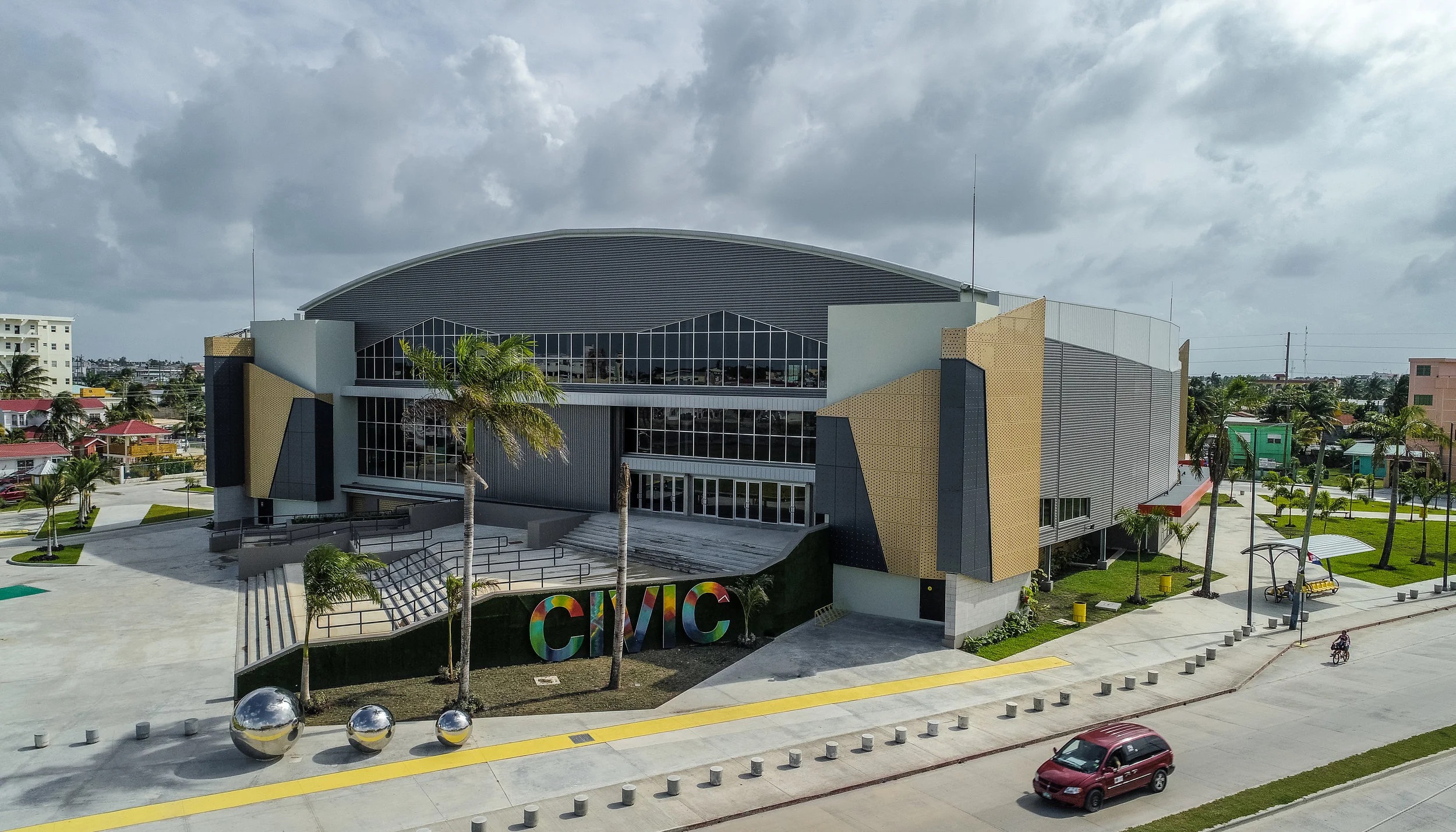 Modern city hall building with large glass windows, surrounded by palm trees, parked cars, and playground equipment, under a cloudy sky.