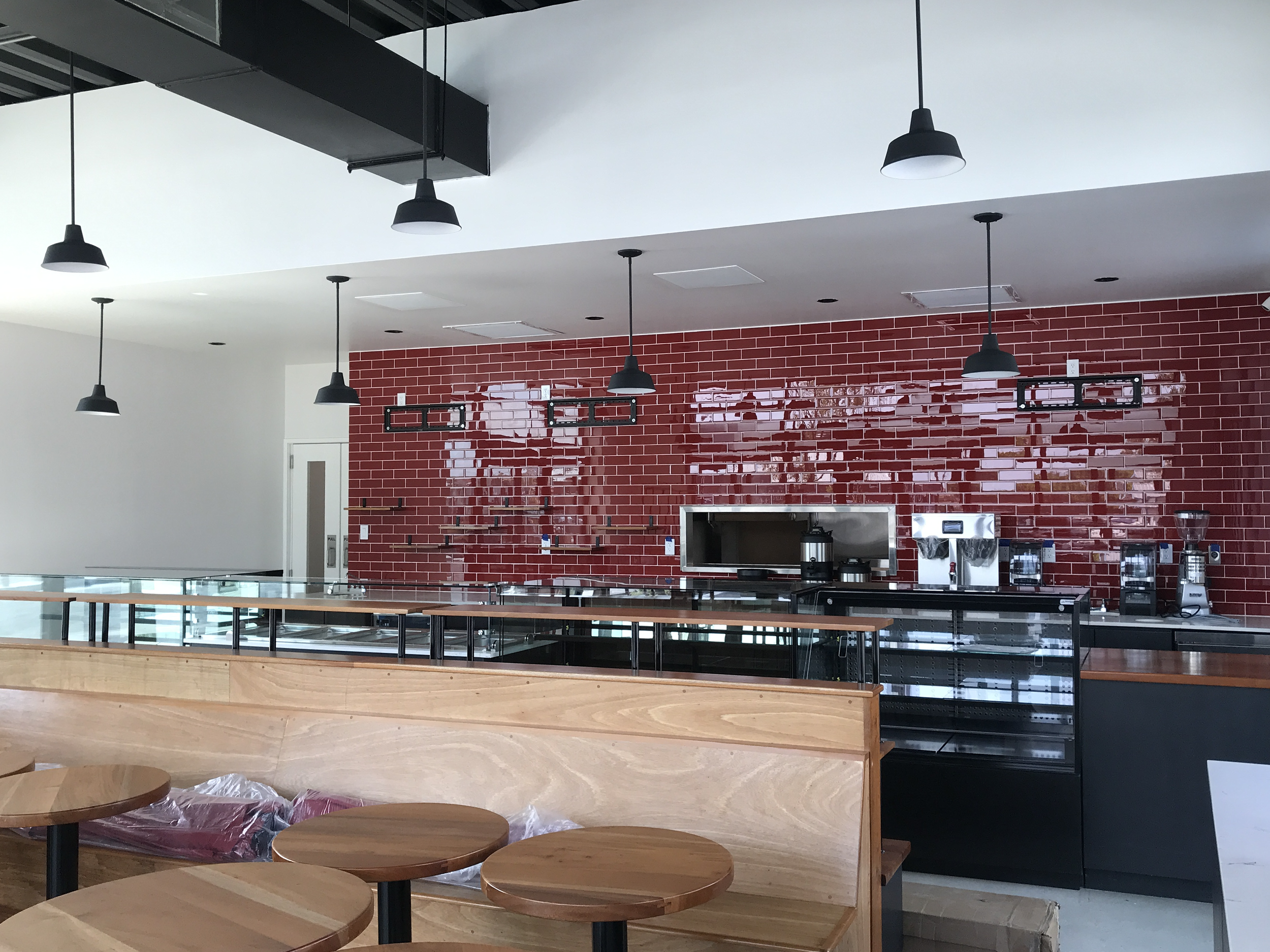 Interior of a modern cafe or restaurant, featuring hanging black pendant lights, a red brick wall, and wooden tables and chairs.