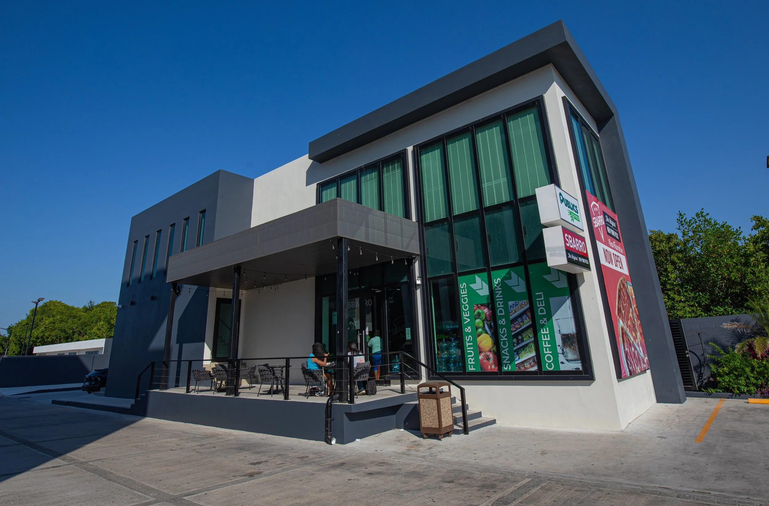 Modern two-story building with large glass windows and a signboard advertising fruits, vegetables, snacks, drinks, coffee, and deli items. There is an outdoor seating area with chairs and tables, and a few people are sitting. The sky is clear and blue, and there are trees and a parking lot in the background.