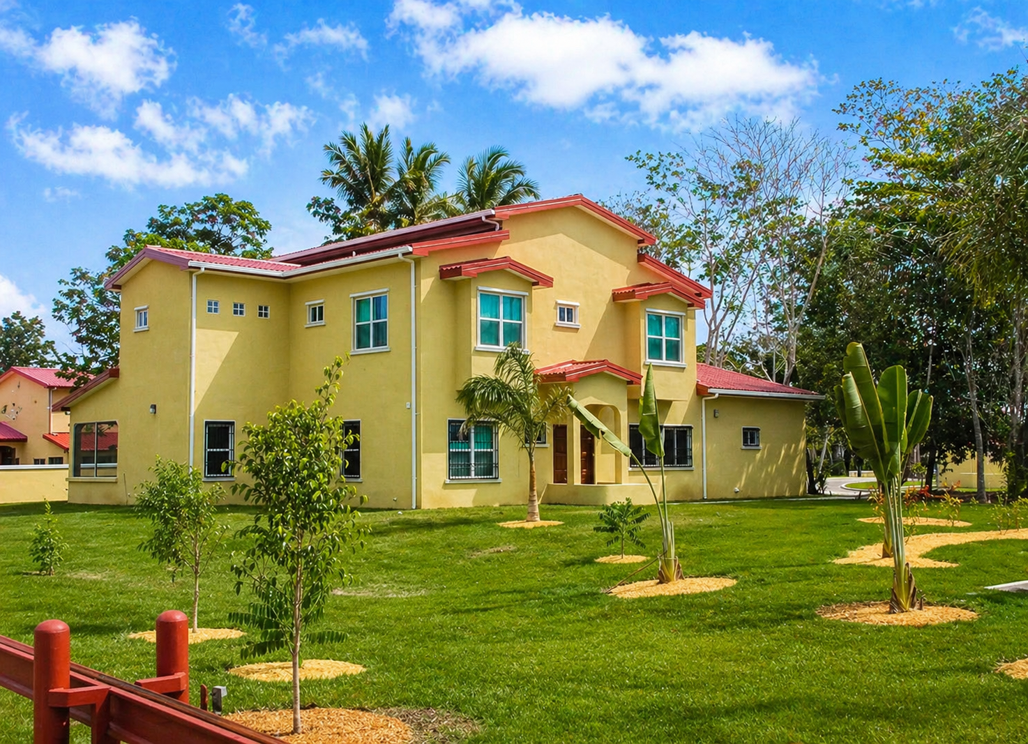 Yellow two-story house with red roof on lush green lawn with small trees and tropical plants, under a partly cloudy blue sky.
