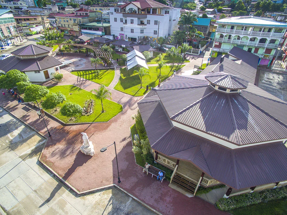 An aerial view of a park surrounded by buildings, with paved walkways, green lawns, trees, and a gazebo, located in an urban area.