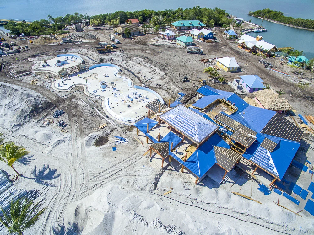 Aerial view of a construction site with a swimming pool and several buildings being constructed near a waterway, with additional small buildings and trees in the background.