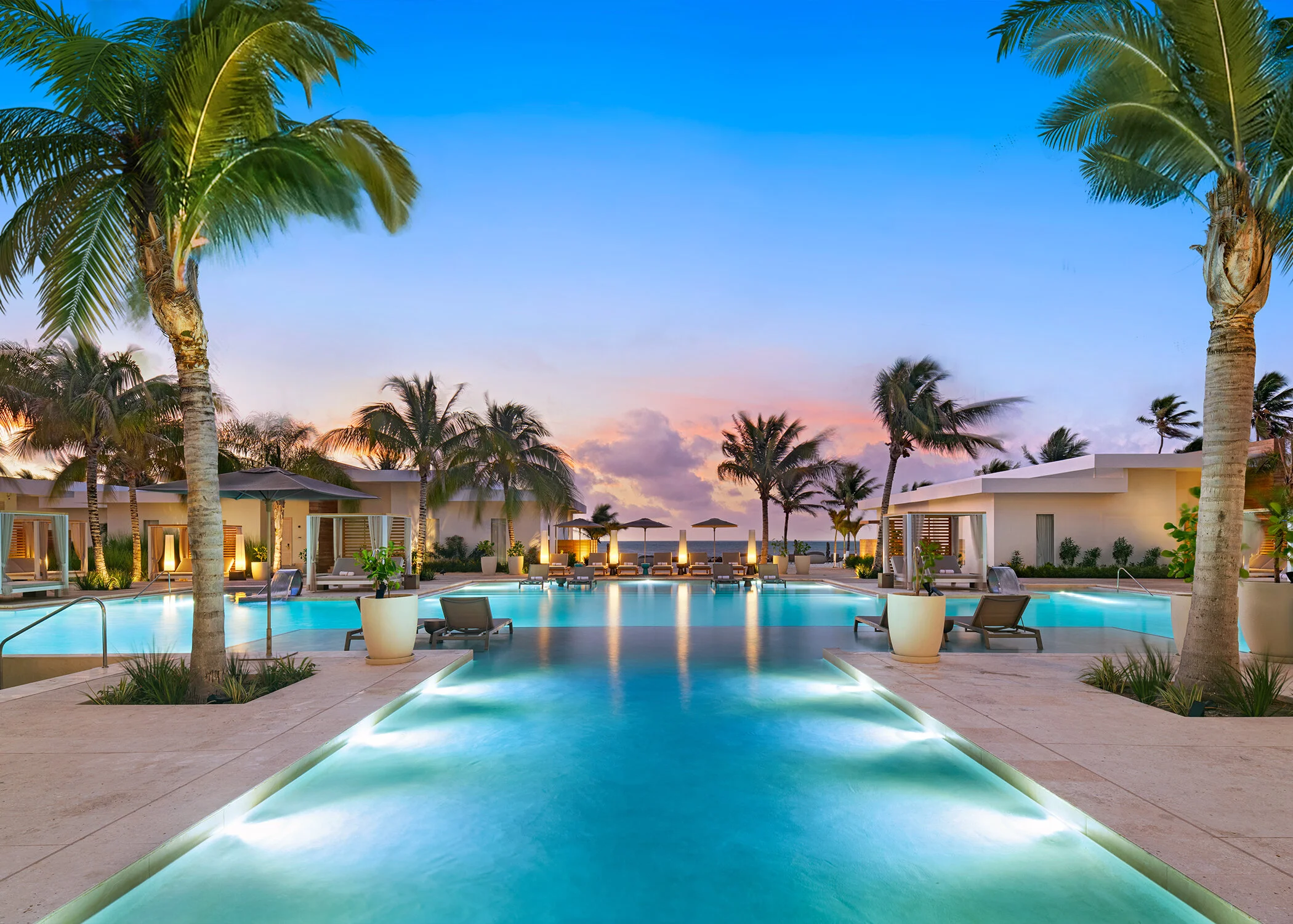 A luxurious pool area at sunset with palm trees, modern lounge chairs, umbrellas, and contemporary buildings, overlooking the ocean