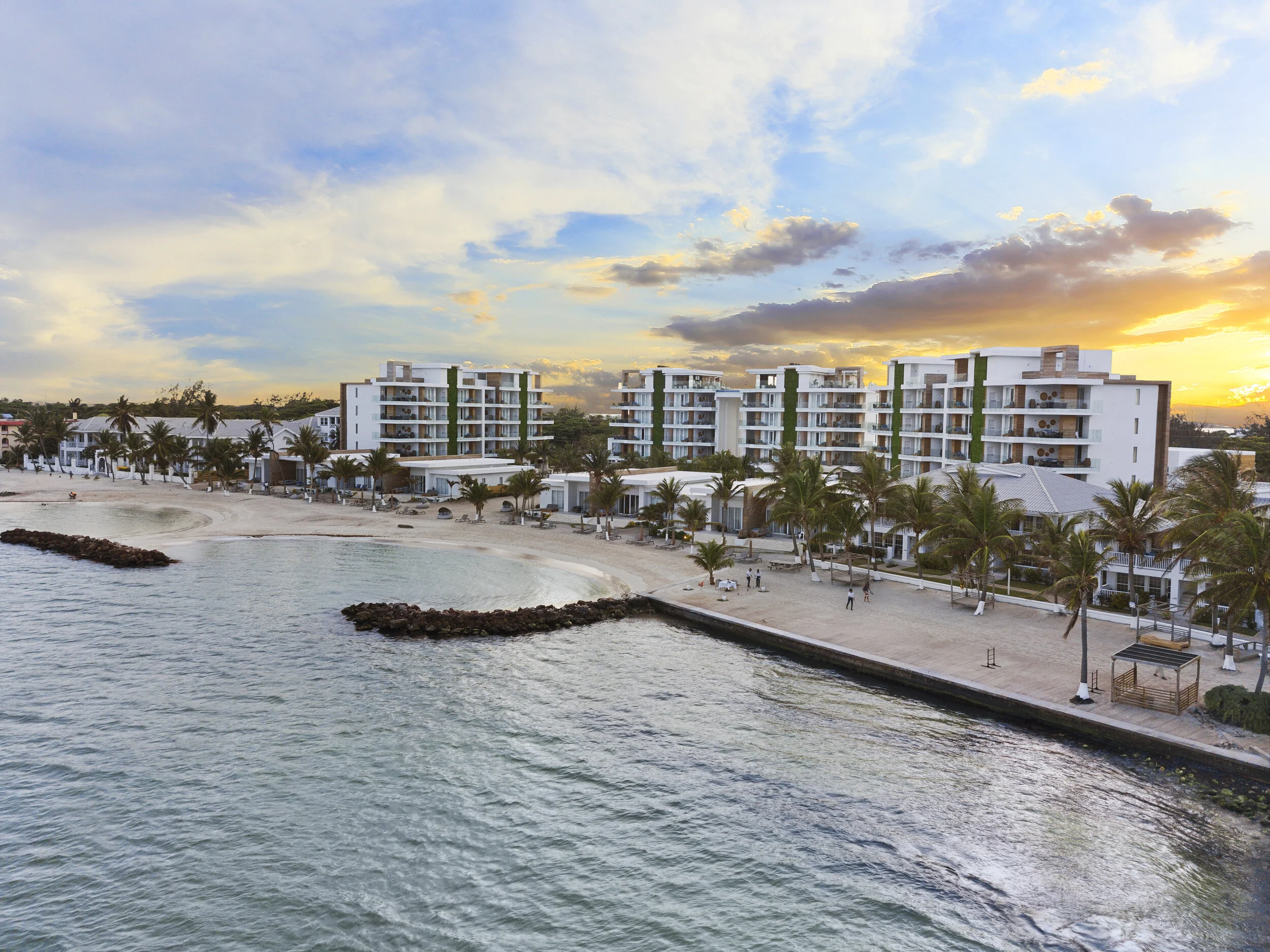 A seaside resort with modern white apartment buildings overlooking the ocean and sandy beach at sunset, with palm trees and people walking on the beach.
