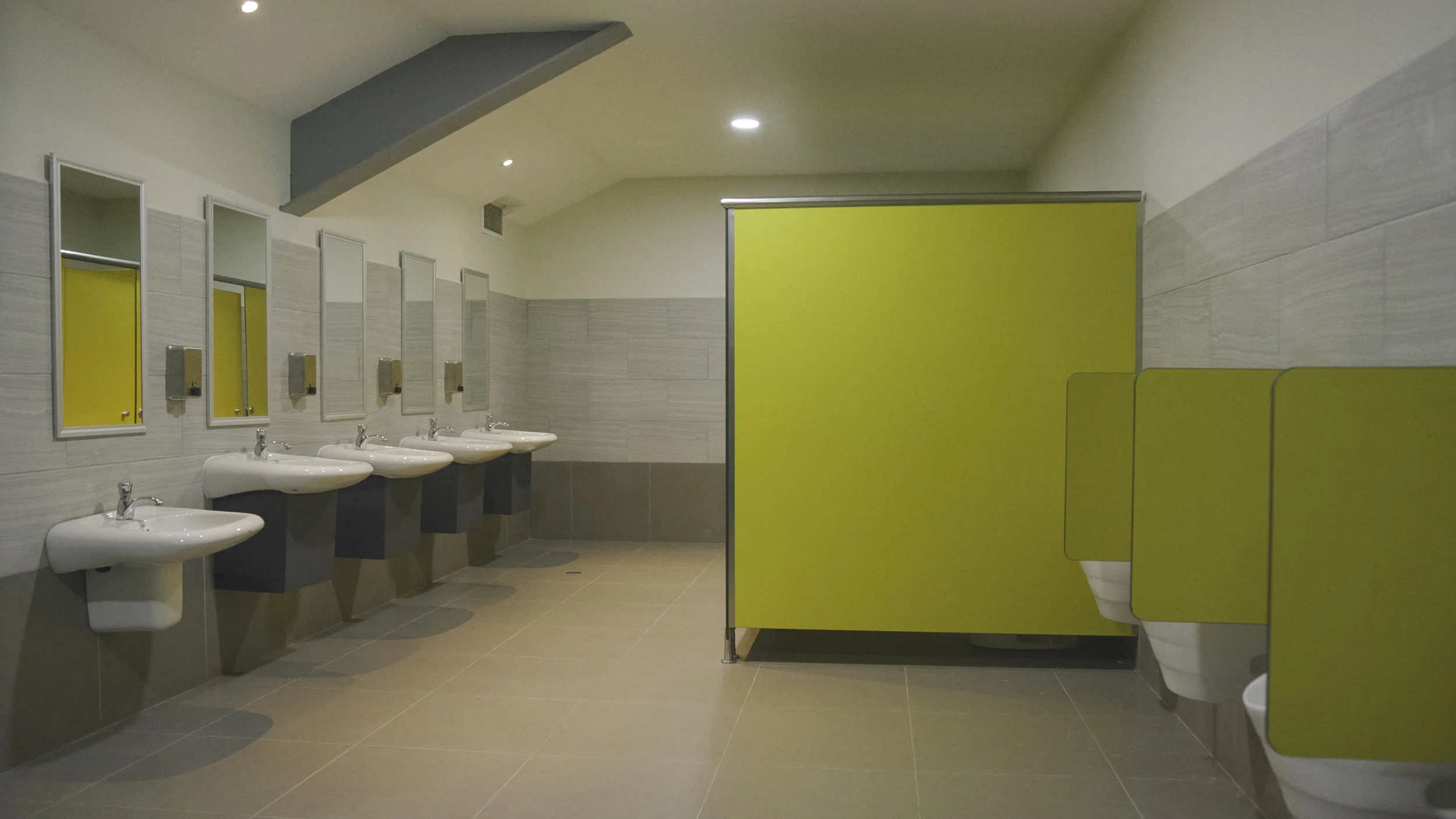 A public restroom with four sinks and mirrors on the wall. To the right, there are green bathroom stalls, including one with a door partially open.