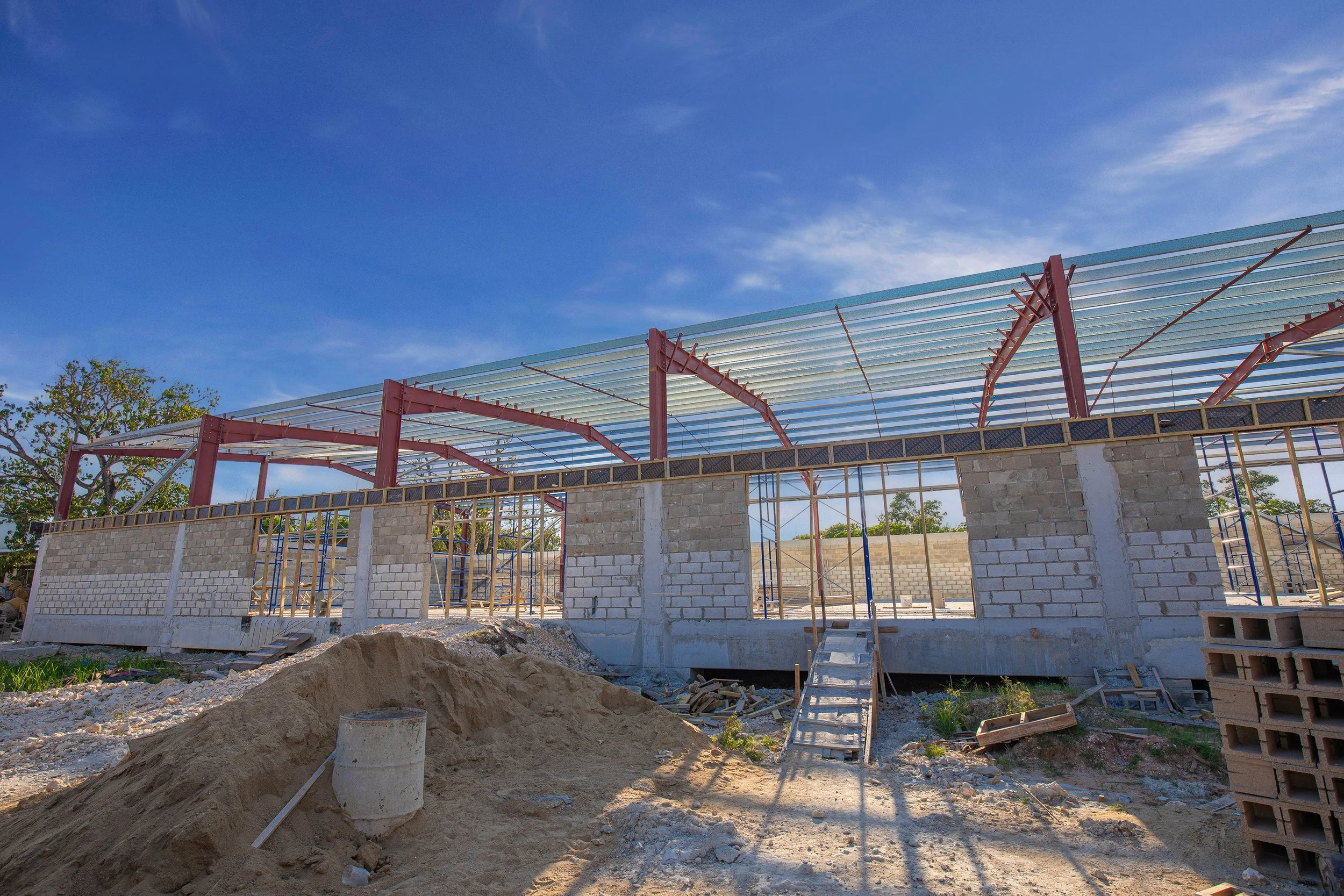 Construction site with a brick building under construction, steel roof framework, scattered construction materials, and a blue sky with some clouds.