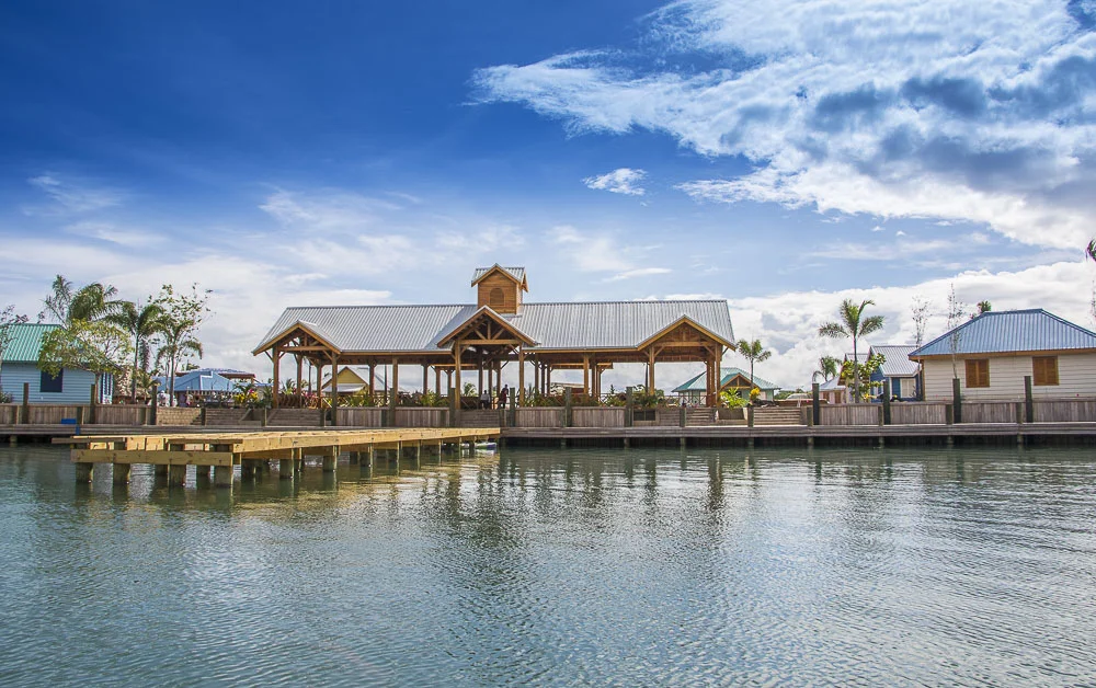 A waterfront scene with wooden buildings and a pier, under a partly cloudy sky, with palm trees and calm water in the foreground.