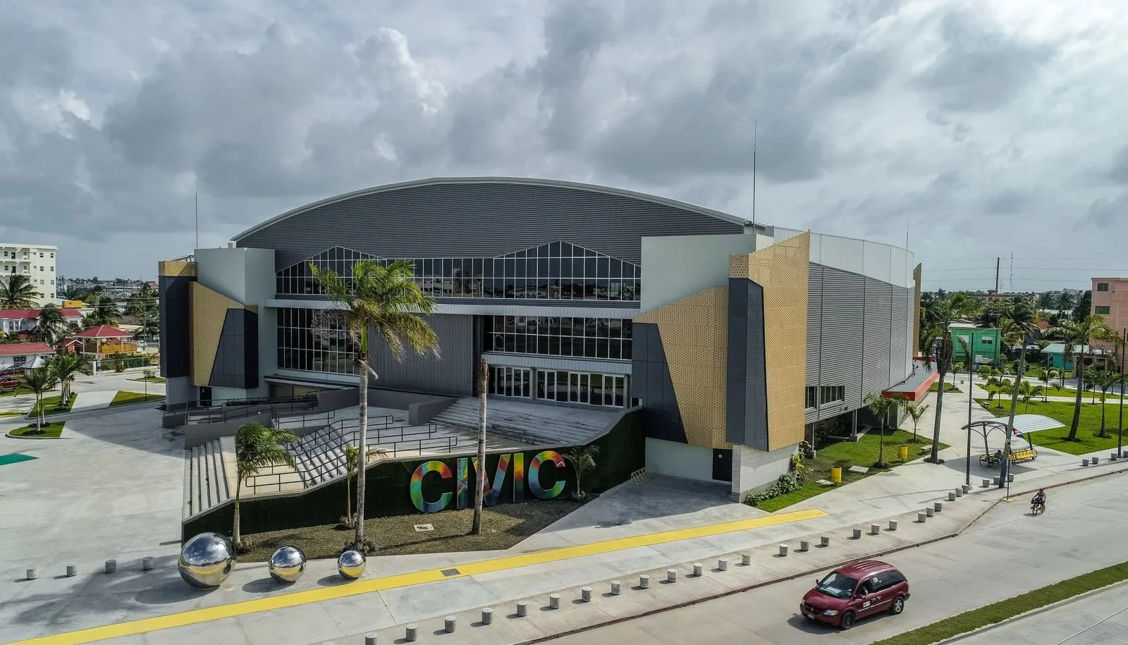 Modern Civic Center building with large stained glass windows, colorful 'CIVIC' sign, palm trees, sidewalk, and street with cars and bicycles, under a cloudy sky.