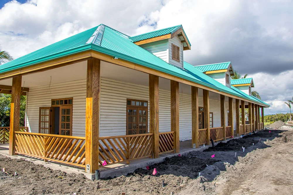 A row of newly built houses with green metal roofs, white siding, and wooden porches. The houses are in a tropical setting with palm trees, and there is ongoing landscaping or construction work in the dirt areas in front.