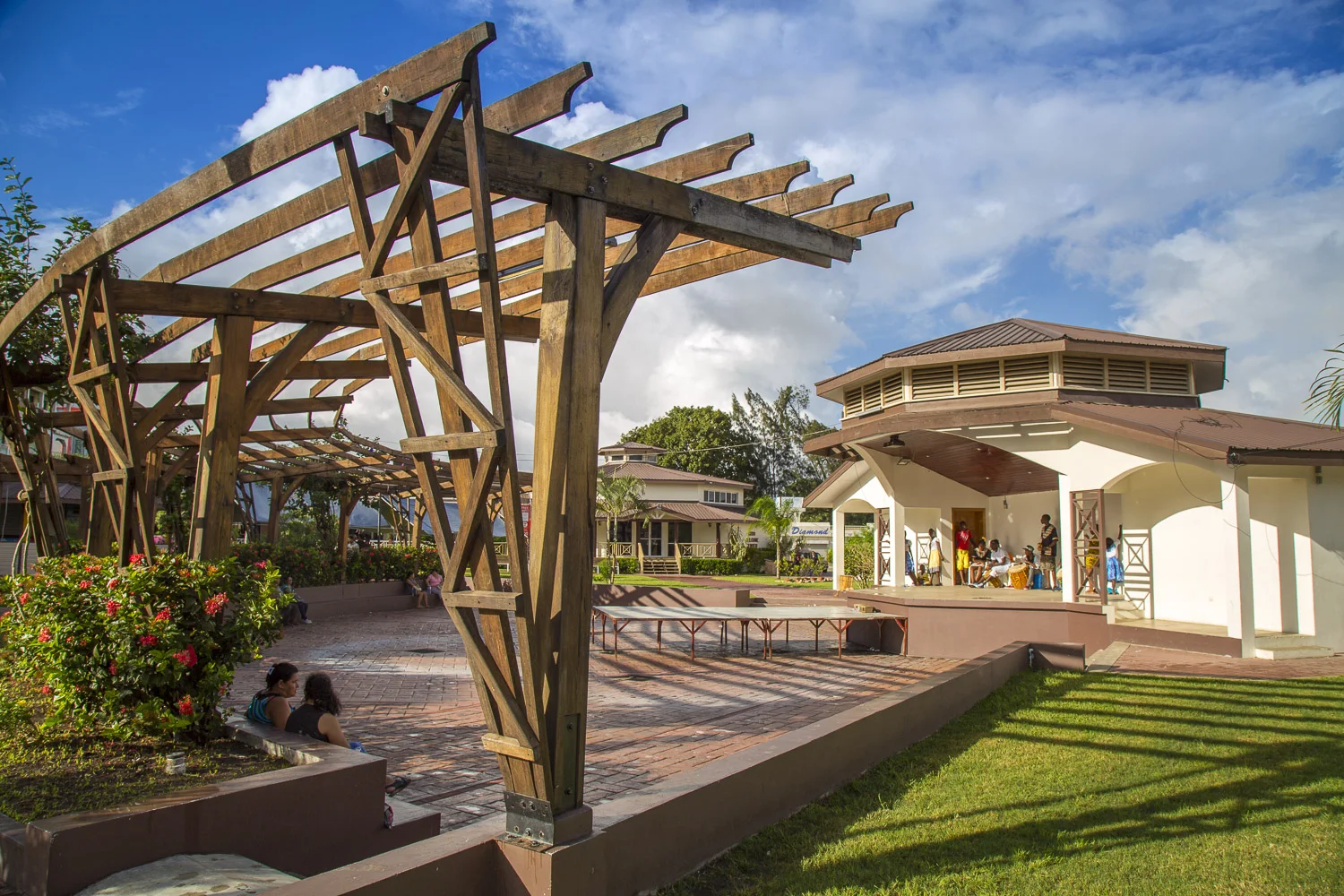 An outdoor community space with a wooden pergola, a small stage, and a white pavilion where people are gathered.