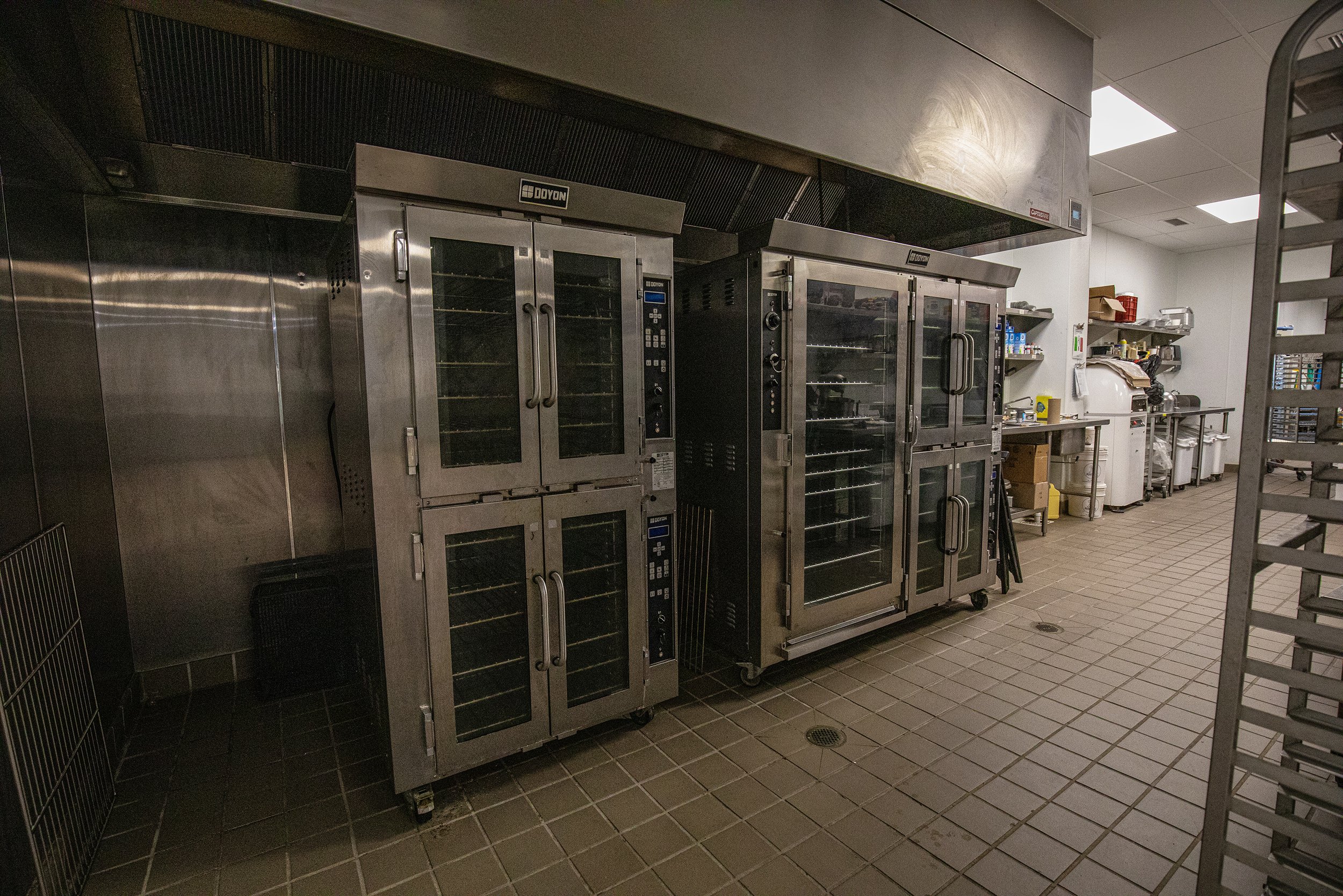 Commercial kitchen with stainless steel appliances, including convection ovens and racks, on a tiled floor, with shelves and equipment in the background.
