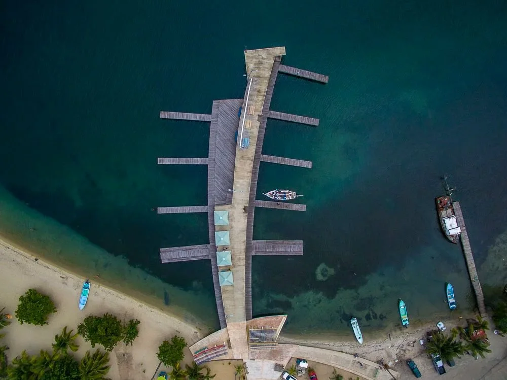 An aerial view of a beach pier with boats docked at the side, surrounded by water and trees along the shoreline.