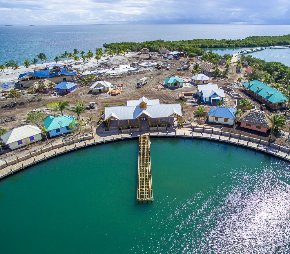 Aerial view of a developing waterfront resort with small colorful buildings, a dock, and a circular lake, surrounded by water and greenery.