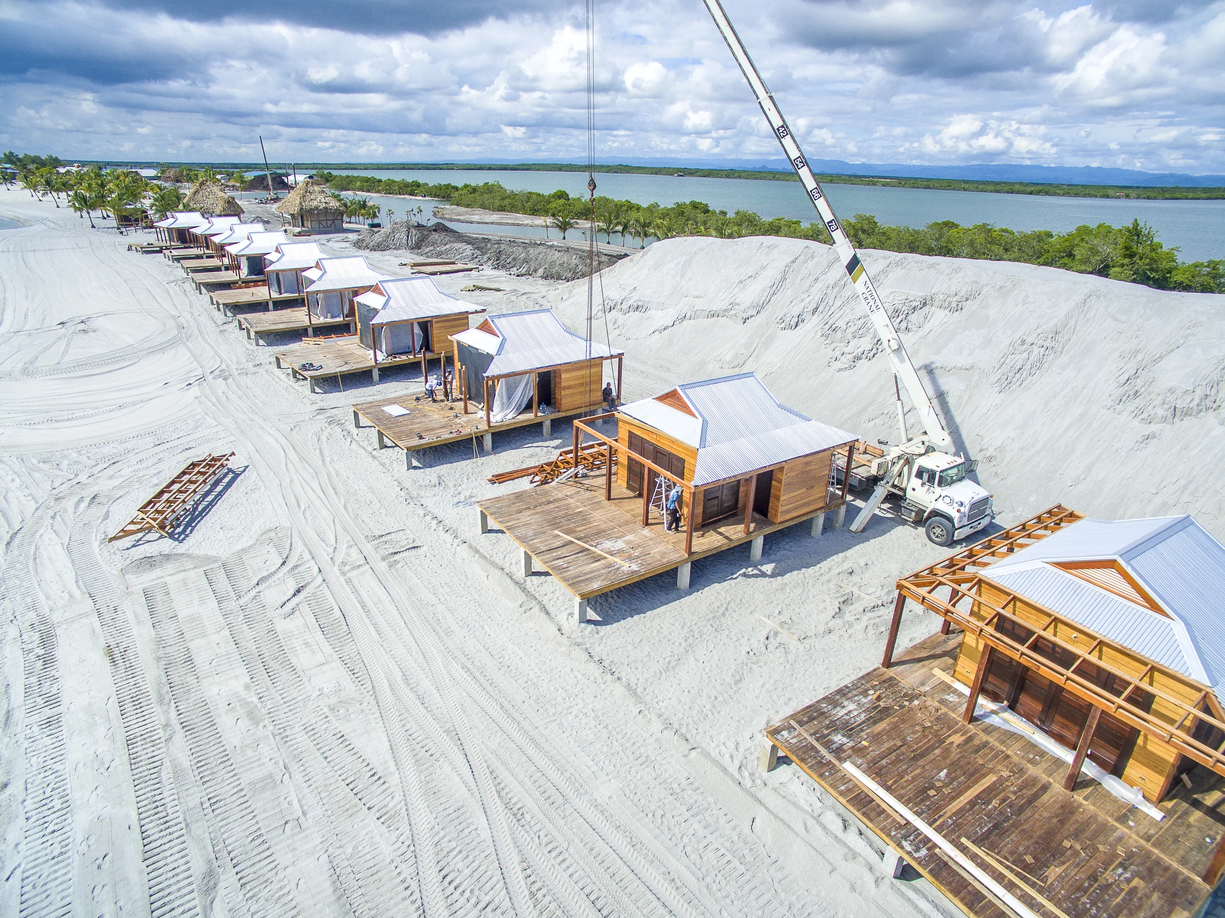 Under construction beachfront bungalows with white roofs, wooden decks, and a crane lifting materials, near a water body with greenery and mountains in the background.